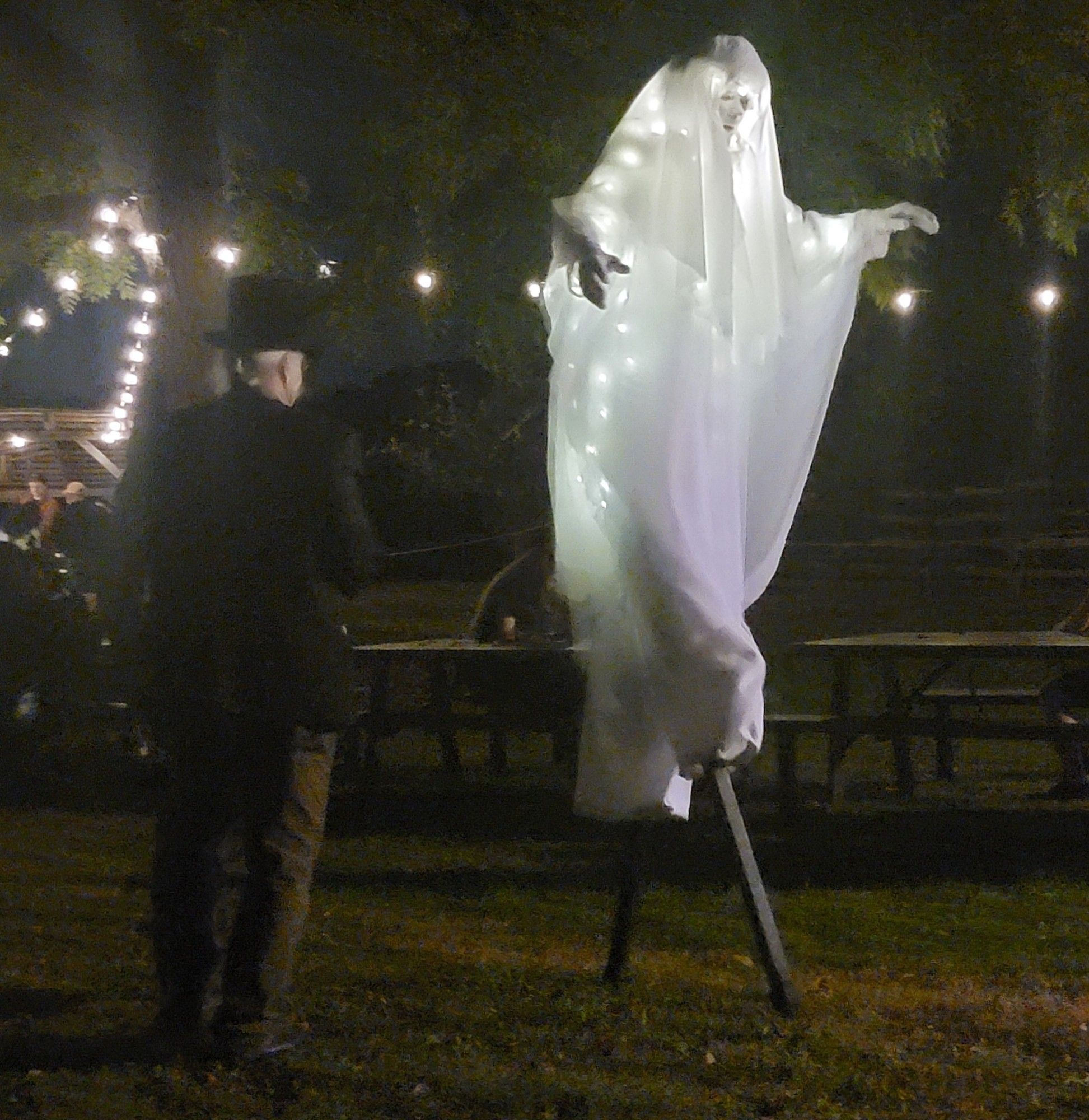 A person facing away from the camera in a tophat beside someone on stilts in gauzey white fabric with lights and a mask that looks like a ghost