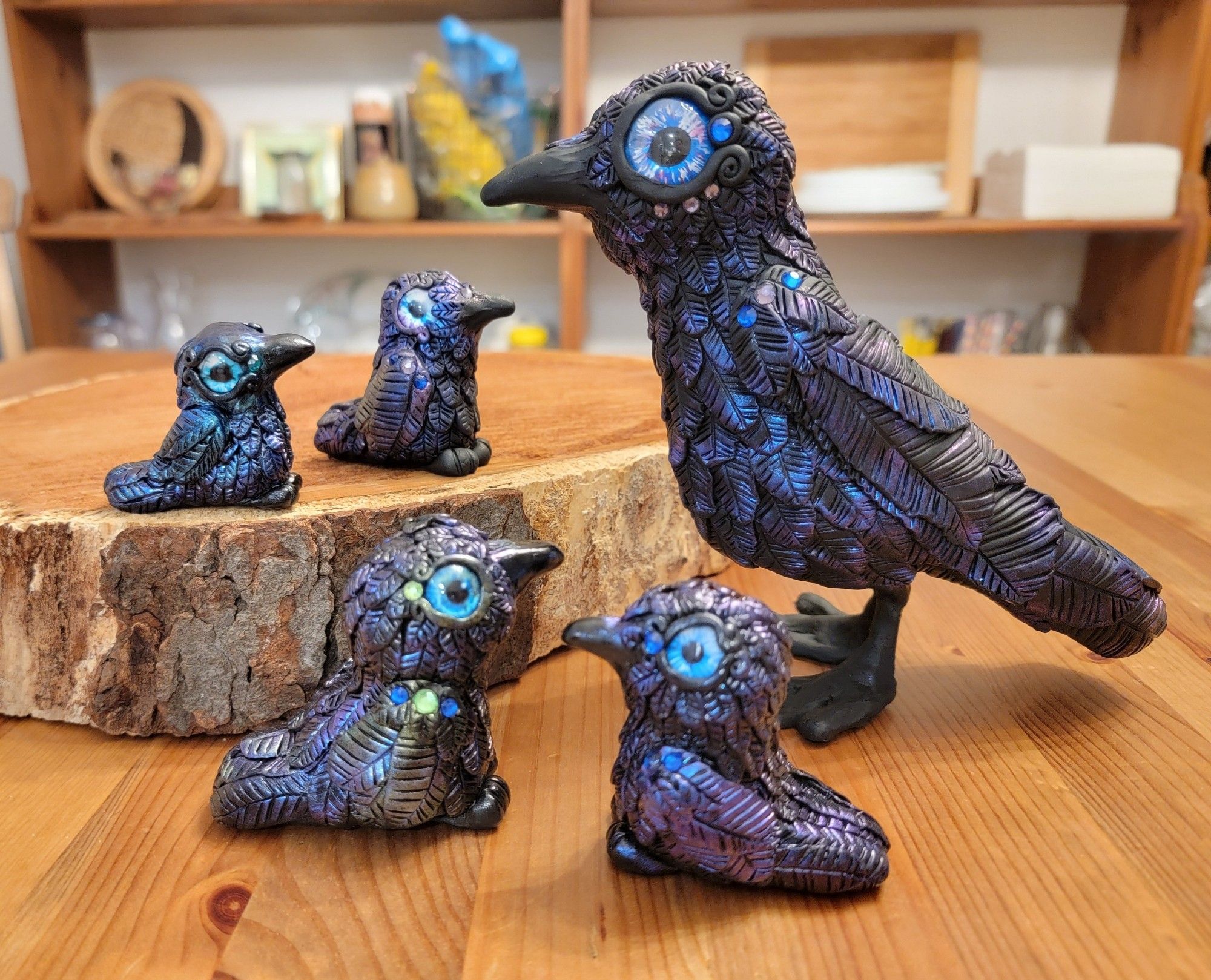 Five polymer clay crows on a table, with glass eyes, shiny feathers, and rhinestone accents.  The smallest is probably under 2 inches tall and the largest is about 6 inches tall.