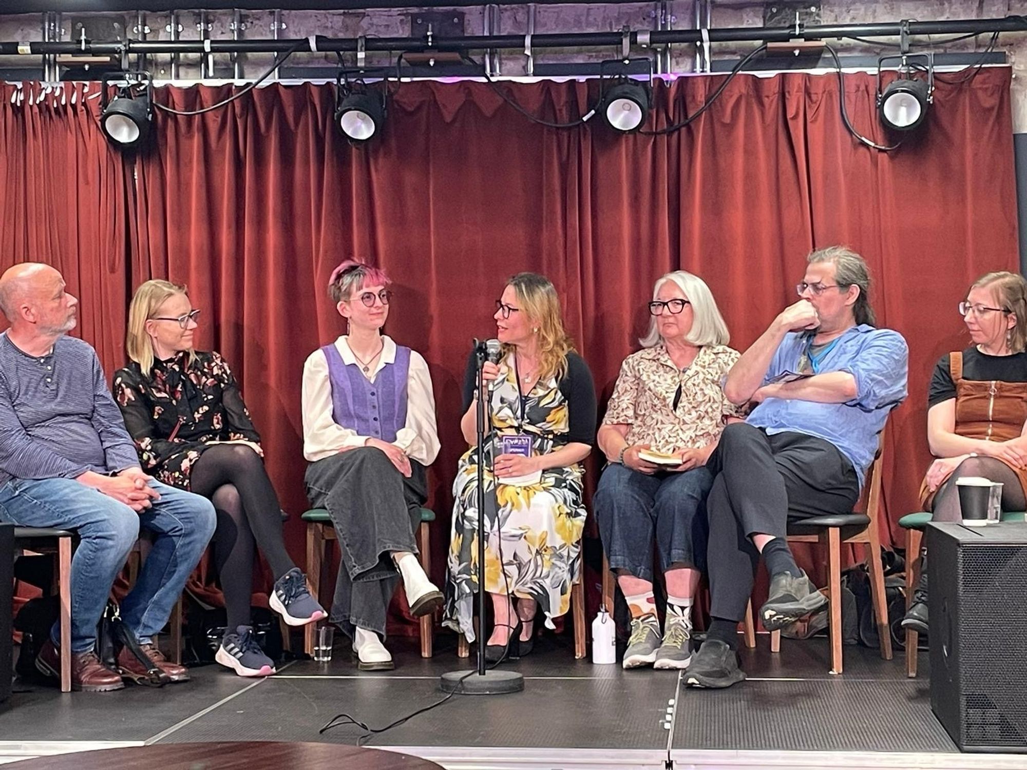 Writers Douglas Thompson, Nadya Mercik, Rayn Epremian, Teika Marija Smits, Anne Charnock, Adrian Tchaikovsky, and Fiona Mossman sit on a stage. Teika is speaking.