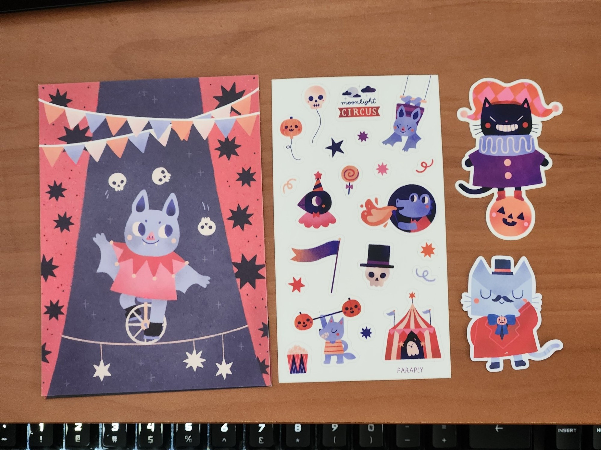 Photo of print and set of stickers themed around a dark circus, featuring cute animals. The print on the left shows a bat riding a unicycle on a tightrope while juggling skulls. The sticker sheet shows the bat on a trapeze, a bird clown, a gator breathing fire, a wolf lifting a barbell with jack-o-lantern weights, a ghost in a big top tent, etc. The two flake stickers are a cat in a harlequin clown outfit balanced on a pumpkin ball, and Lelo the cat in the ring master outfit