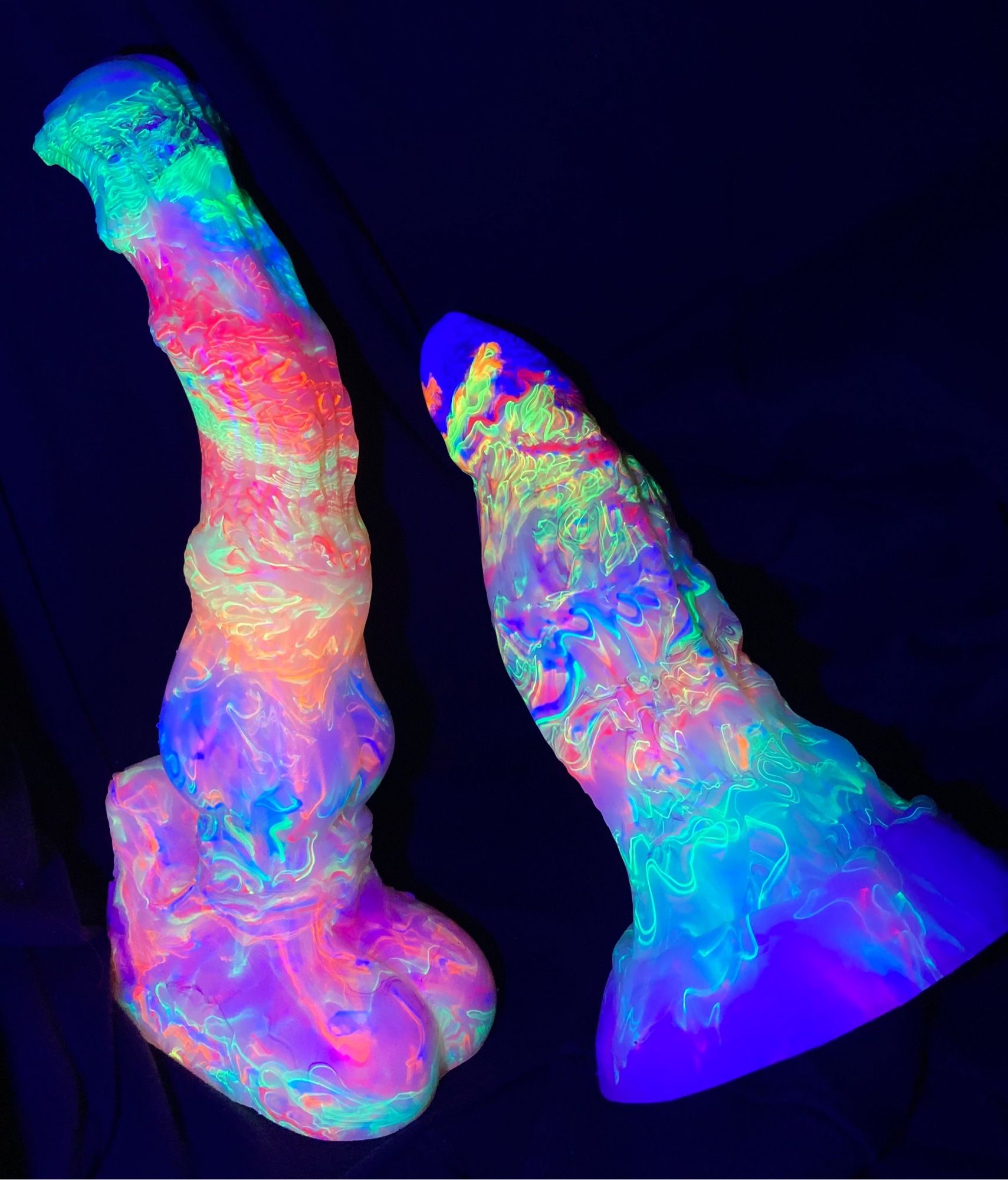 seraphina [left]; pegasus inspired dildo with a flared head, rippling shaft and know

tempestt [right]; a highly textured thick dragon inspired dildo with a significant swell

both toys are illuminated under UV light and the rainbow colors of the toys show as scattered bright ribbons