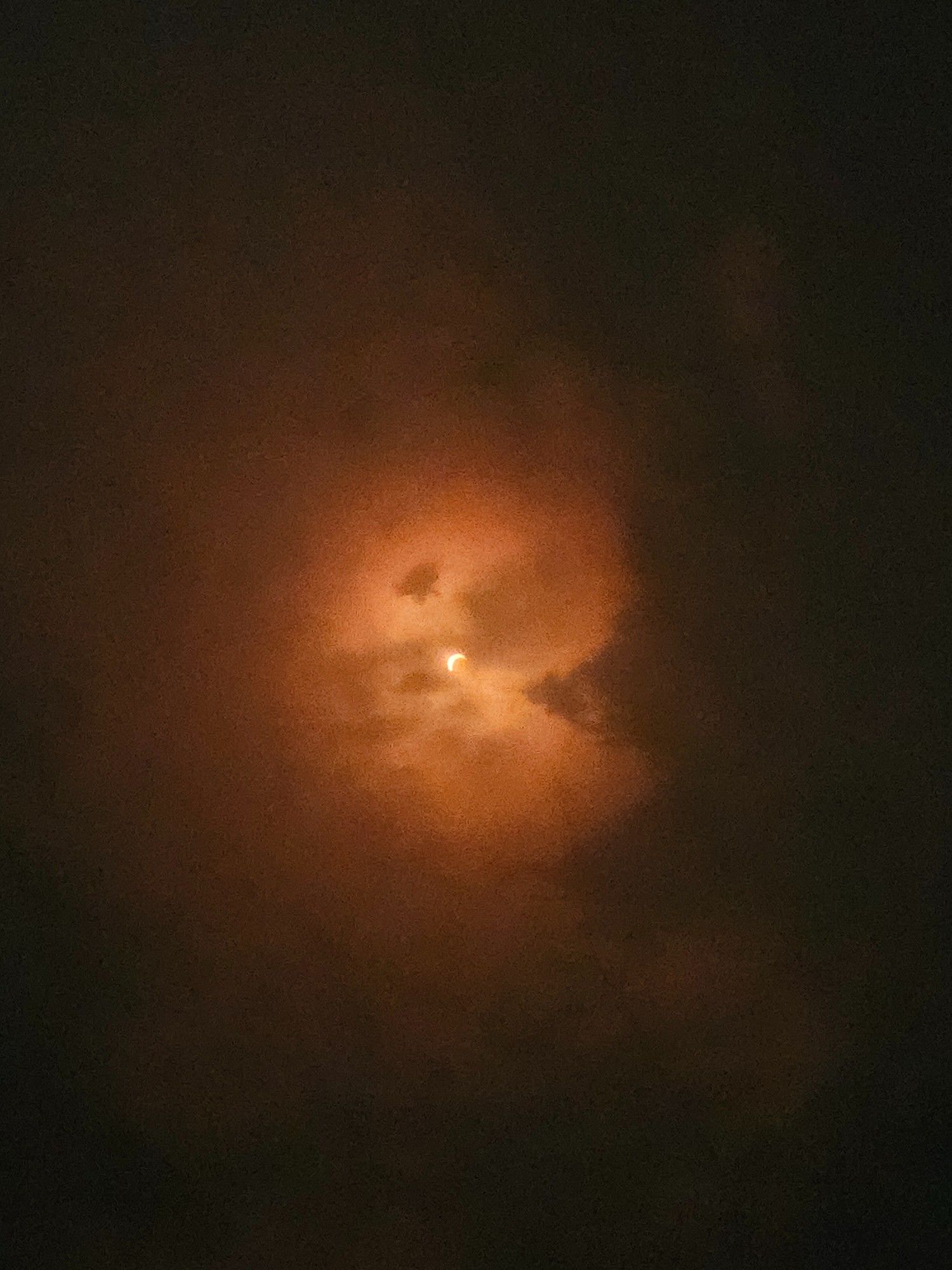 a photo of a solar eclipse with approximately 80% of the sun obscured by the moon, with the crescent of the sun lighting up clouds around it in an orange glow