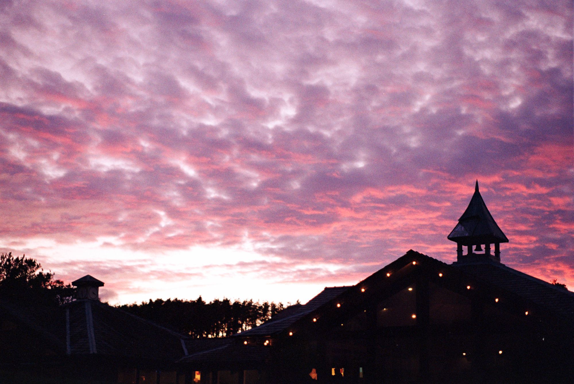 A silhouette of a rest stop against a sky illuminated by a summer sunset, casting orange across the patchy clouds. A group of trees sits between two halves of the building in the composition, also silhouetted against the bright setting sun.