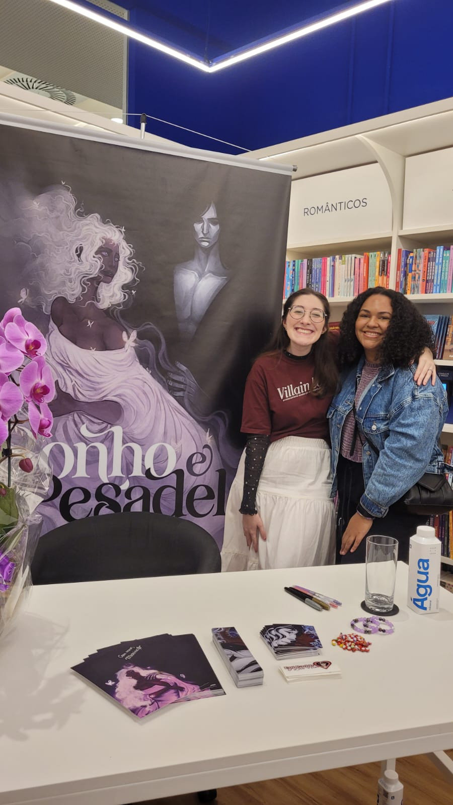 Foto. No fundo, à esquerda, banner com a capa do livro Sonho e Pesadelo. Em uma mesa branca à frente, uma orquídea roxa, caras e marcadores do livro, canetas, pulseiras da amizade, joaninha, um copo de vidro e água. Entre a mesa e o banner, uma cadeira preta, e à direita, Marina Dutra e Karoline Melo abraçadas de lado.