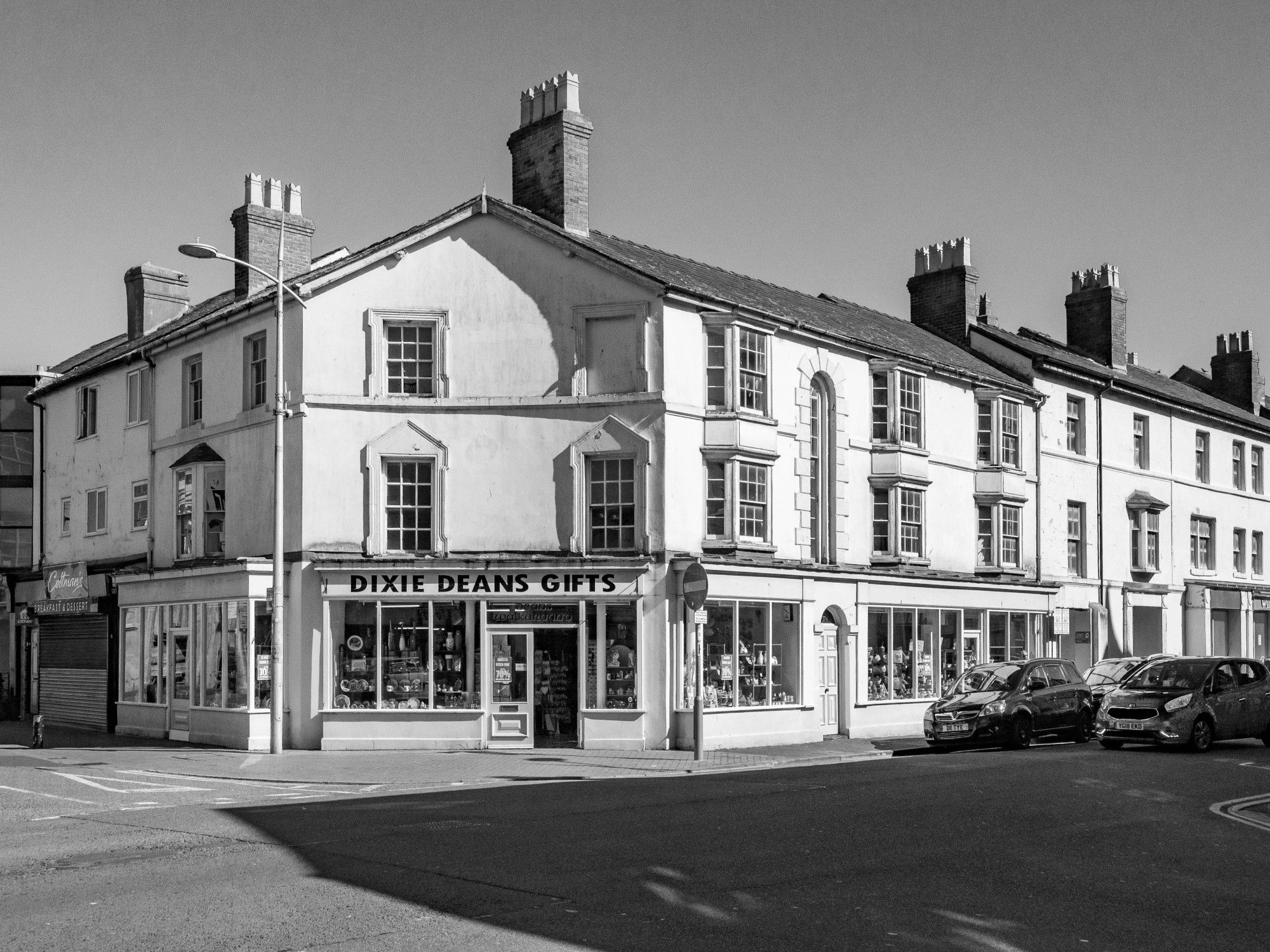 A monochrome image of a street corner in Rhyl with terraced shops, probably built in the 1920s or 1930s. The corner of the terrace is cut off, with a shop front reading "Dixie Deans Gifts".