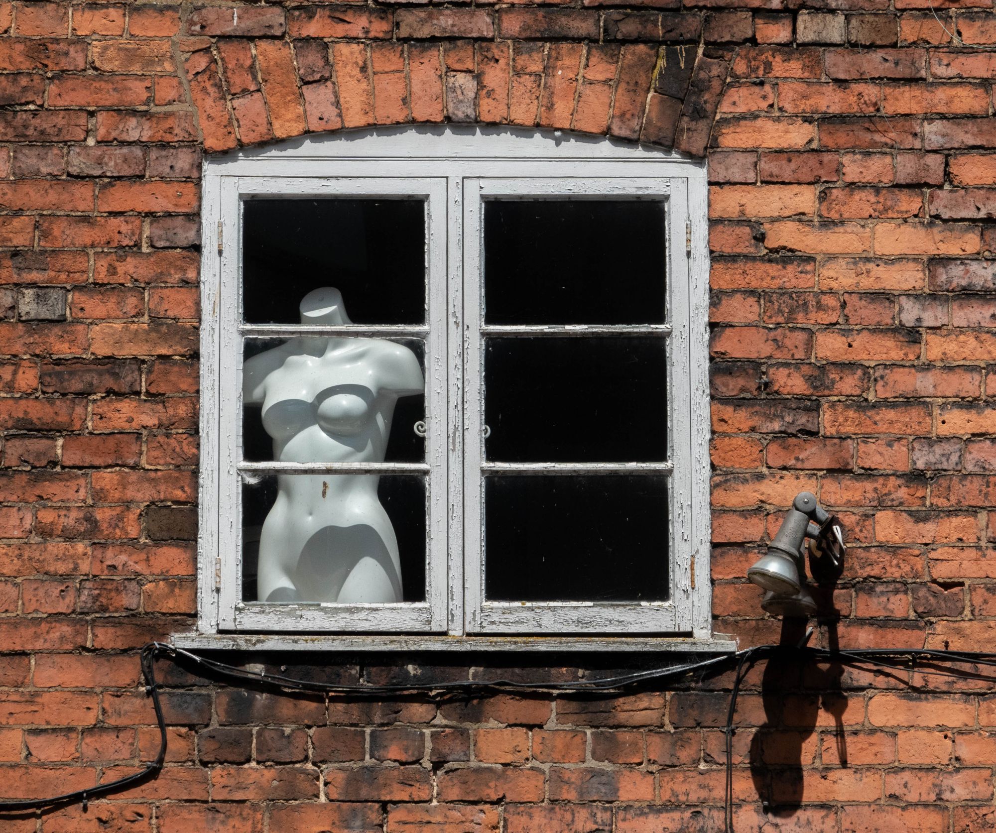 Colour image of a window set in a red-brick wall, with a single shop mannequin of a nude female torso.