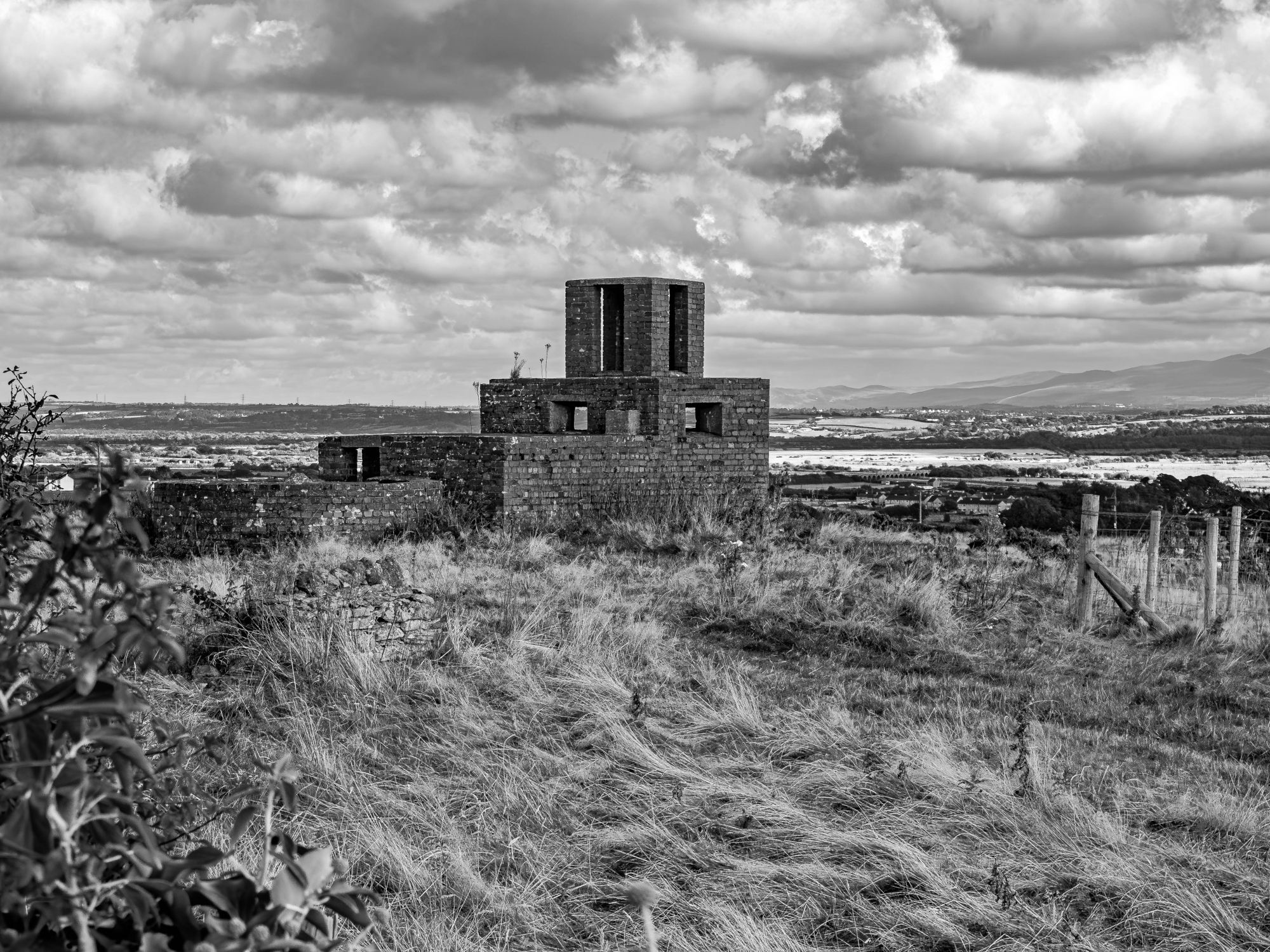 Monochrome image of an unusual, very square abandoned and derelict brick building with 3 distinct levels, in a field with long grass in the foreground and a sky with well-defined clouds. It doesn't conform to a standard farm building's shape, and has the appearance, perhaps, of being a military buiding.