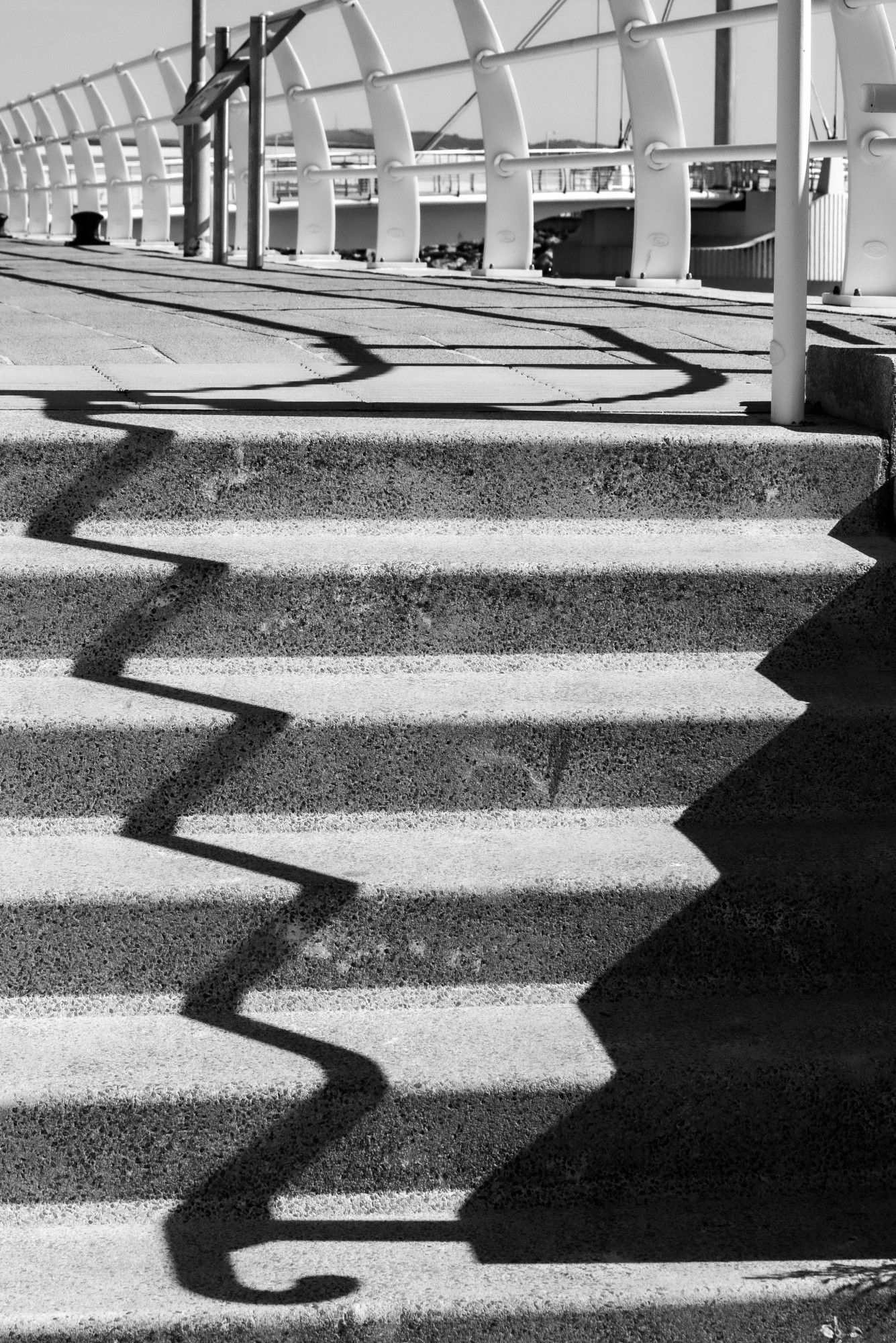 A monochrome image with a flight of six concrete steps, with bright sunshine casting a shadow of railings, out of shot by the steps, but visible in the background at the top of the steps, in a zig-zag pattern on the steps.