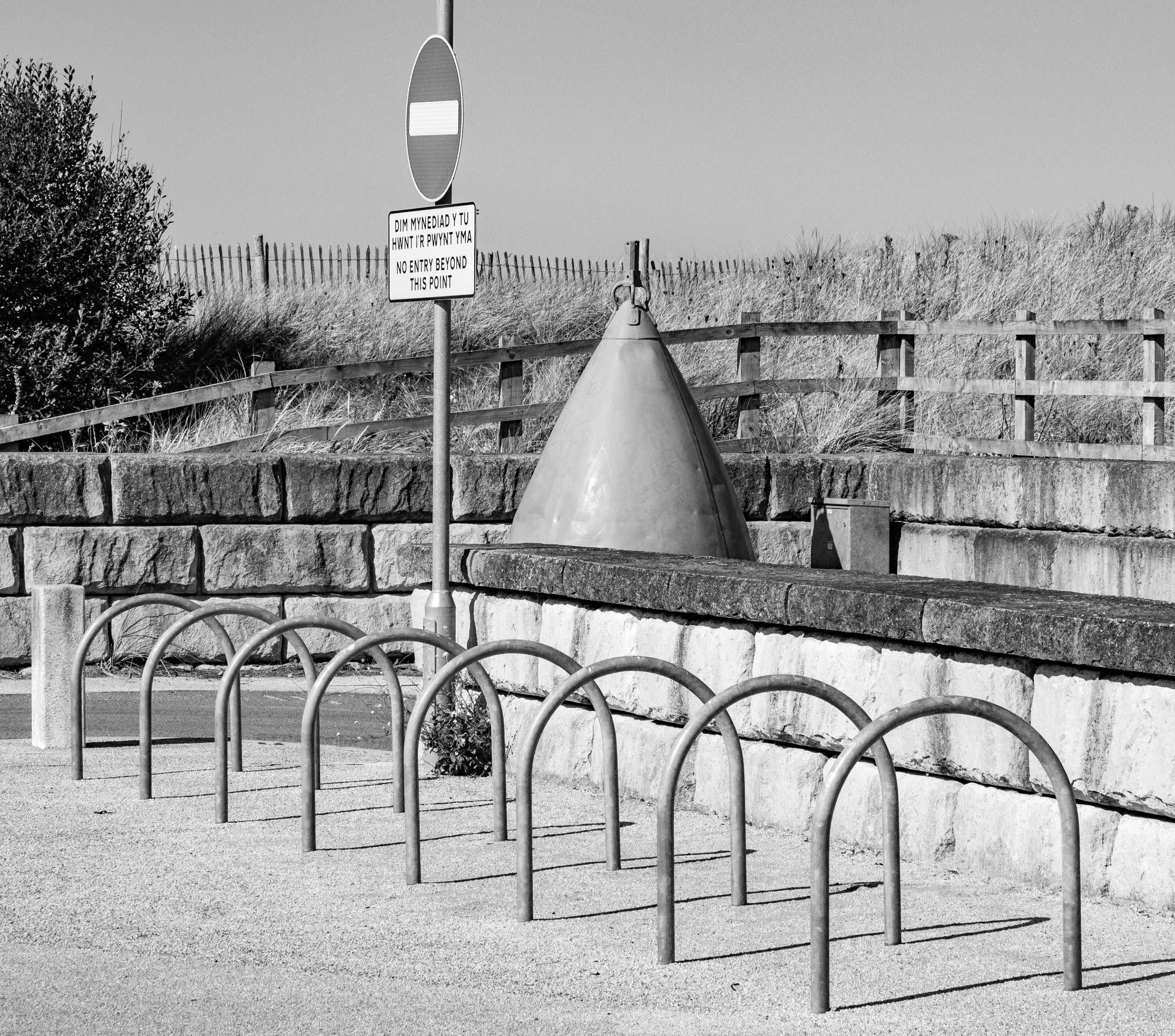 A monochrome image of a buoy in the background, enclosed by a low wall, with a row of hooped bicycle tethering posts, shown in a diagonal line, so that they overlap in a graphic shape.