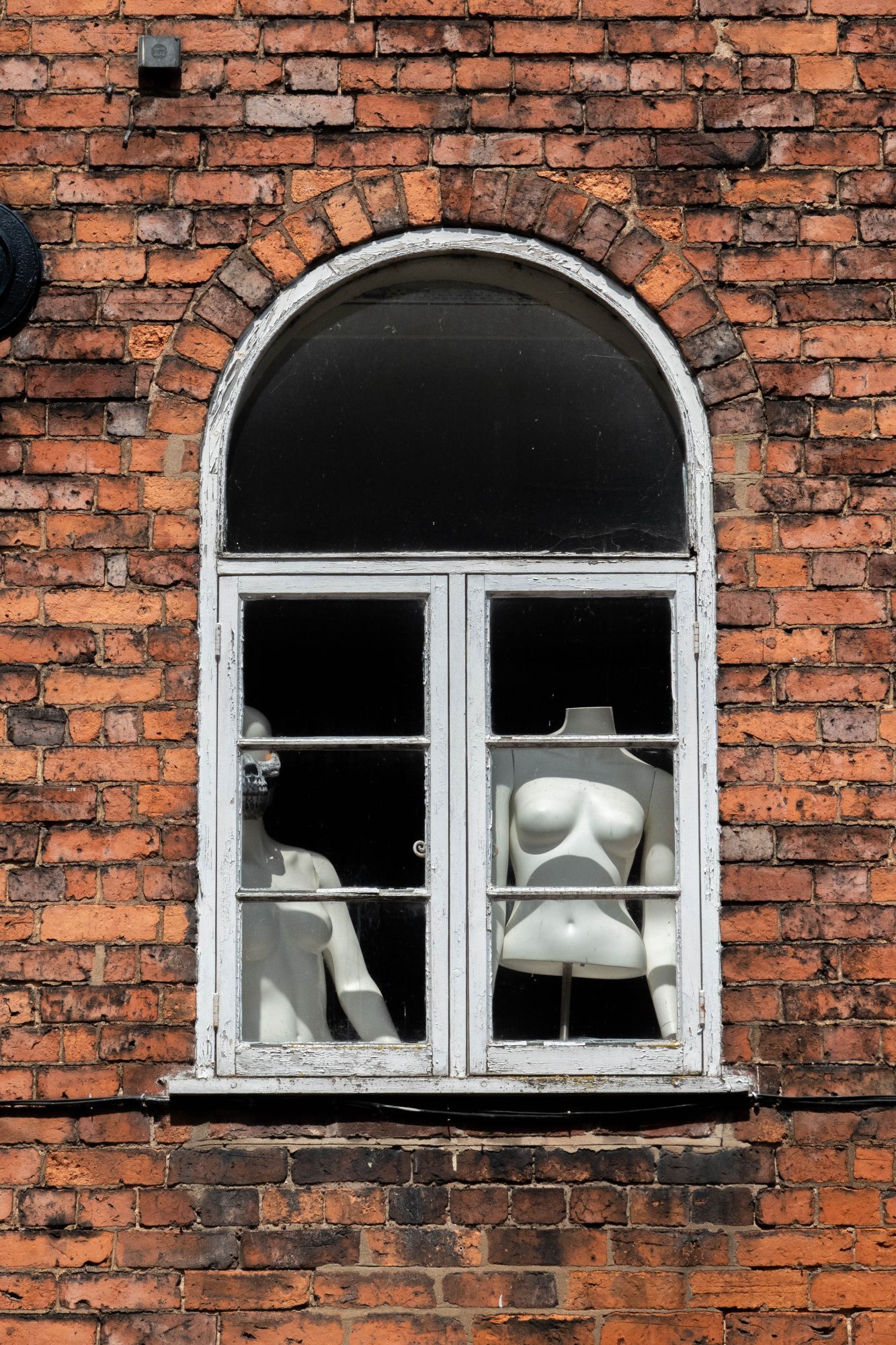 Colour image of a window set in a red-brick wall, with two shop mannequins, one partially hidden, of nude female torsos.