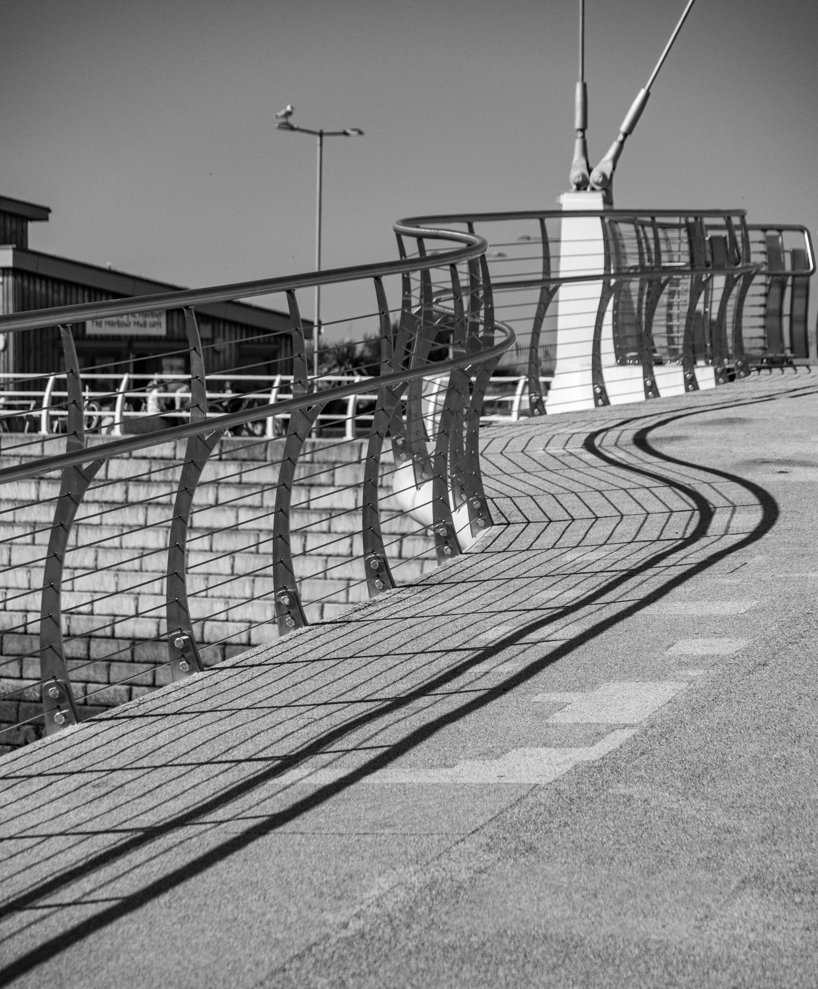 A monochrome image of a curved railing leading to a footbridge (out of shot), with strong shadows projected from the railing onto the footpath.