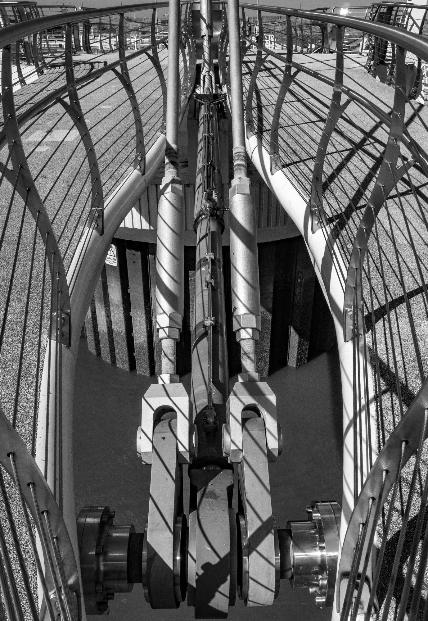 A monochrome image of the anchorings of two metal suspension bridge supports rising from below the bridge footpath, which is divided allowing the supports to be visible.