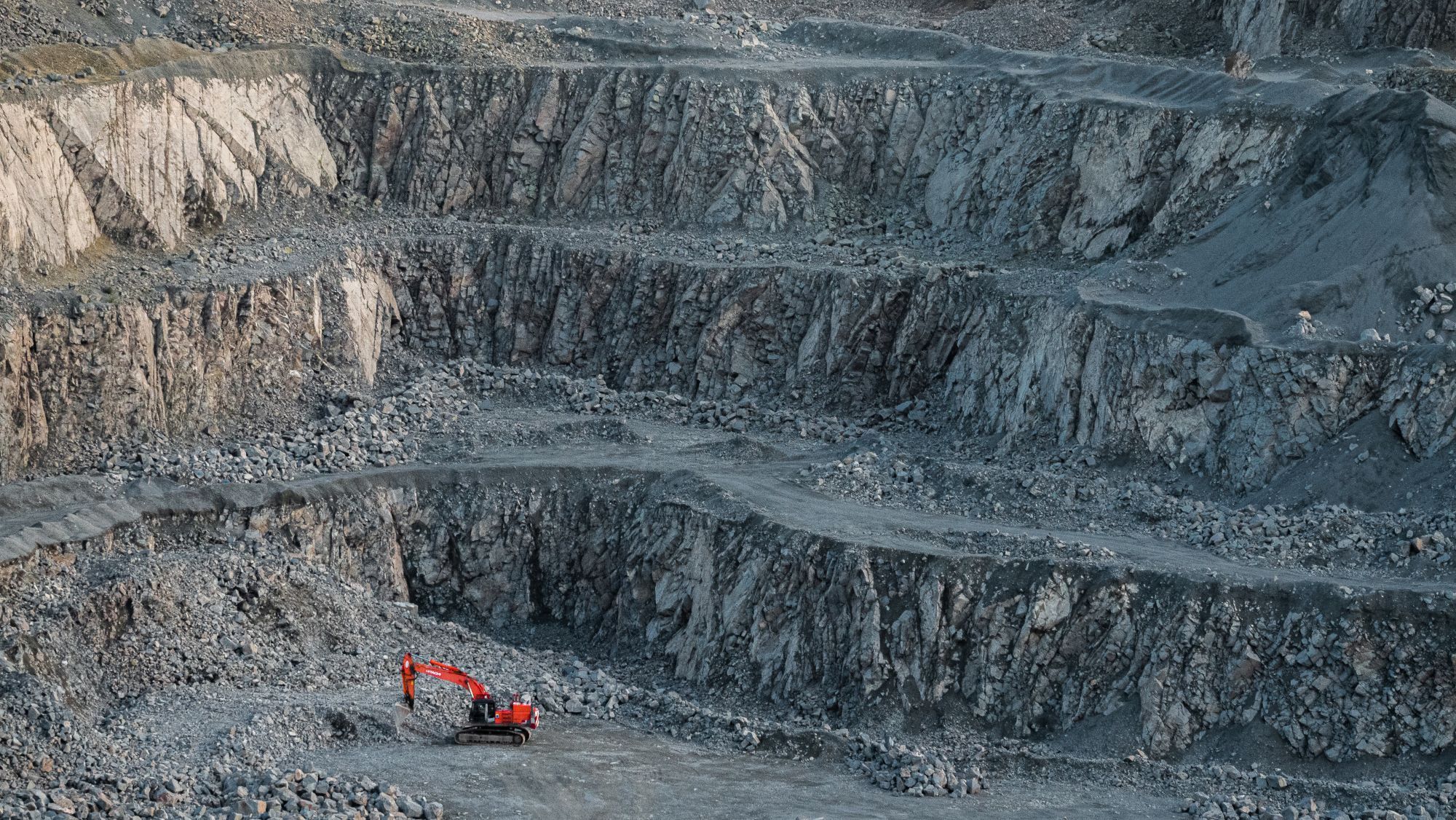 A granite quarry, showing three terraced layers of excavation, with a red digger machine in the bottom left corner of the frame, at the bottom of the pit.