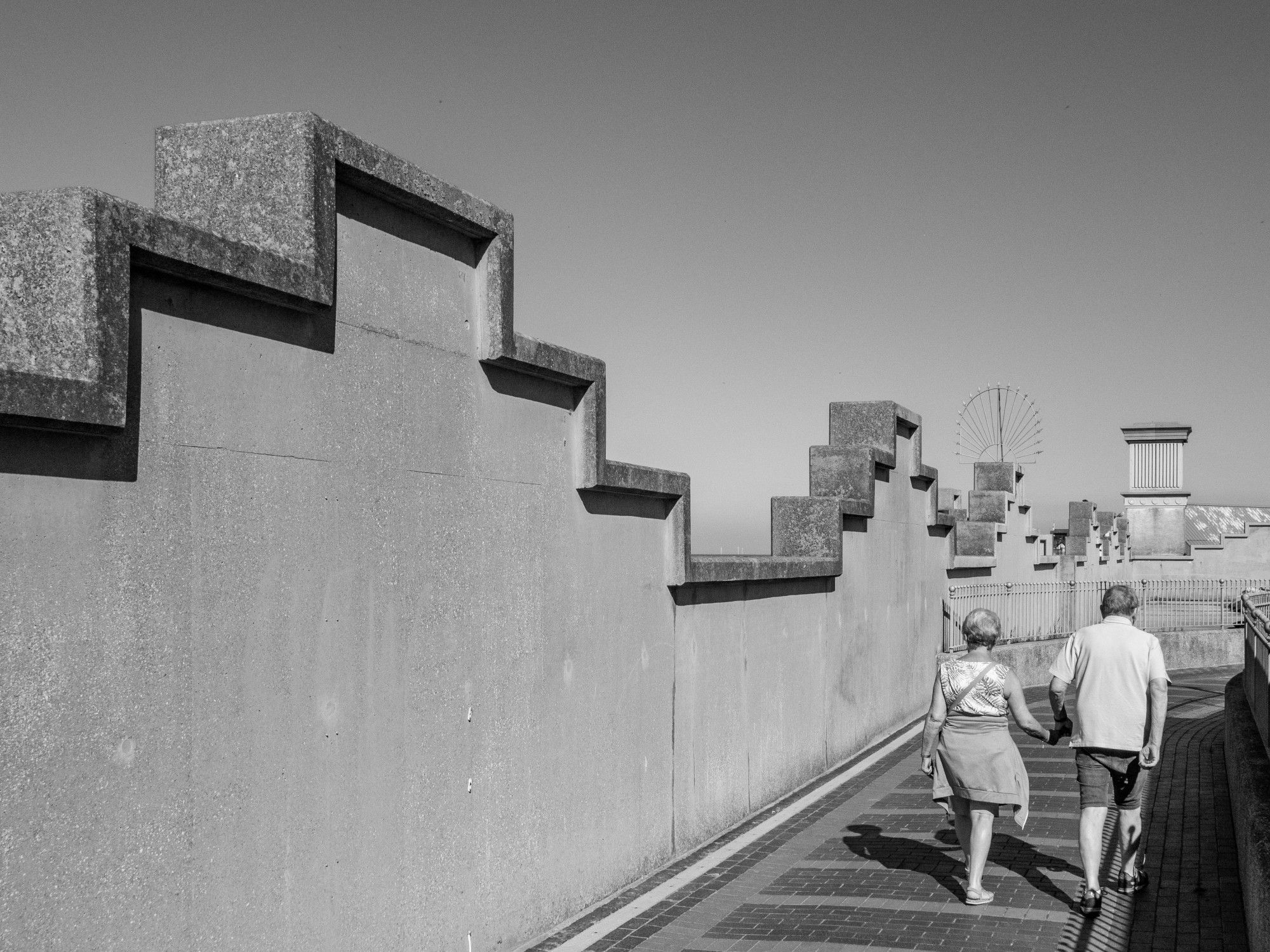 A monochrome image of the rear view of a mature couple walking hand-in-hand alongside a castellated wall.