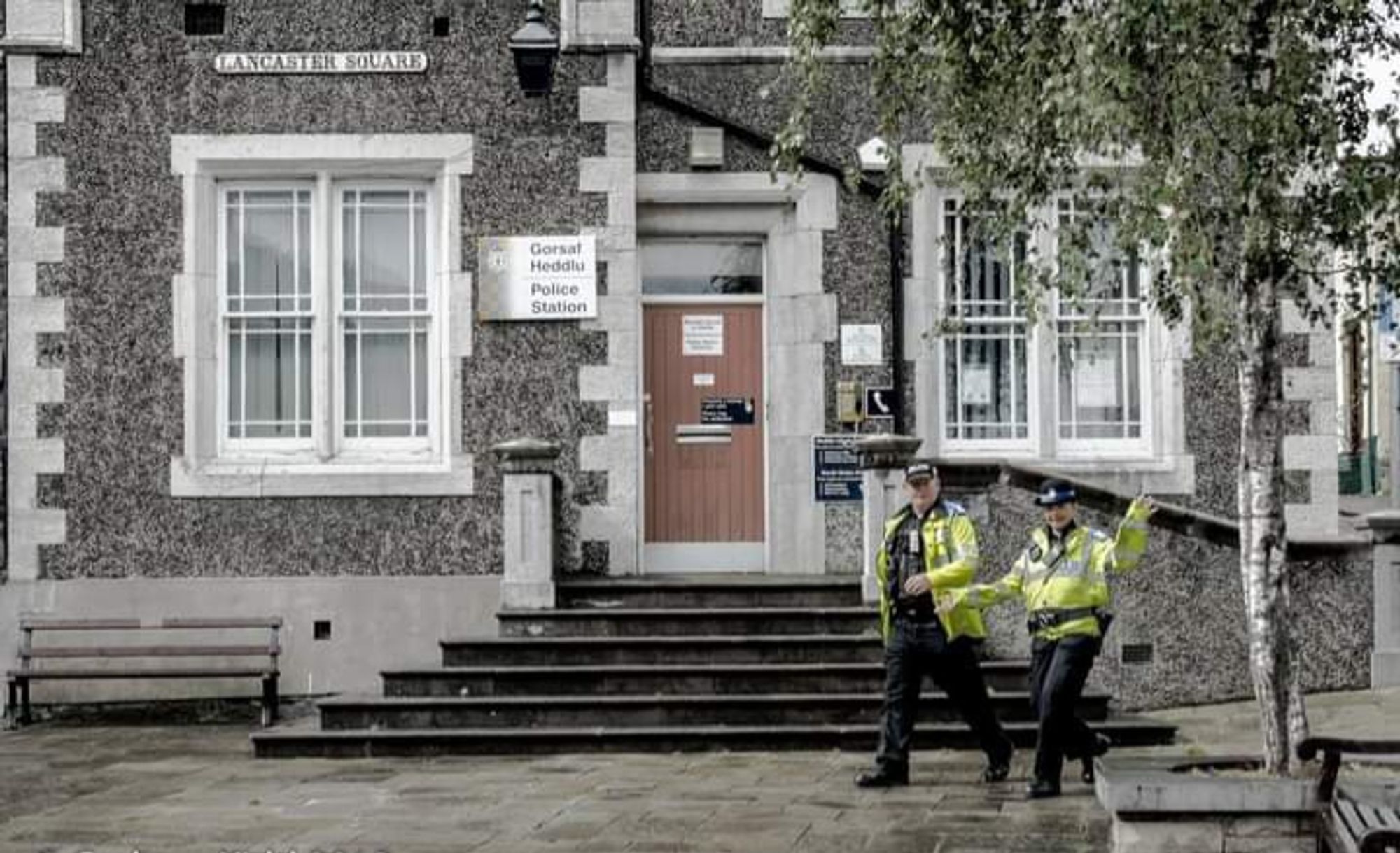 Colour image of the entrance to the Victorian Police station in Lancaster Square, Conwy, with six steps leading to the front door, and sash windows on either side. There's a tree on the left of the frame, with two Police officers, one male & one female,in yellow hi-vis jackets walking into shot. They are smiling, and the female office is giving me "jazz hands".