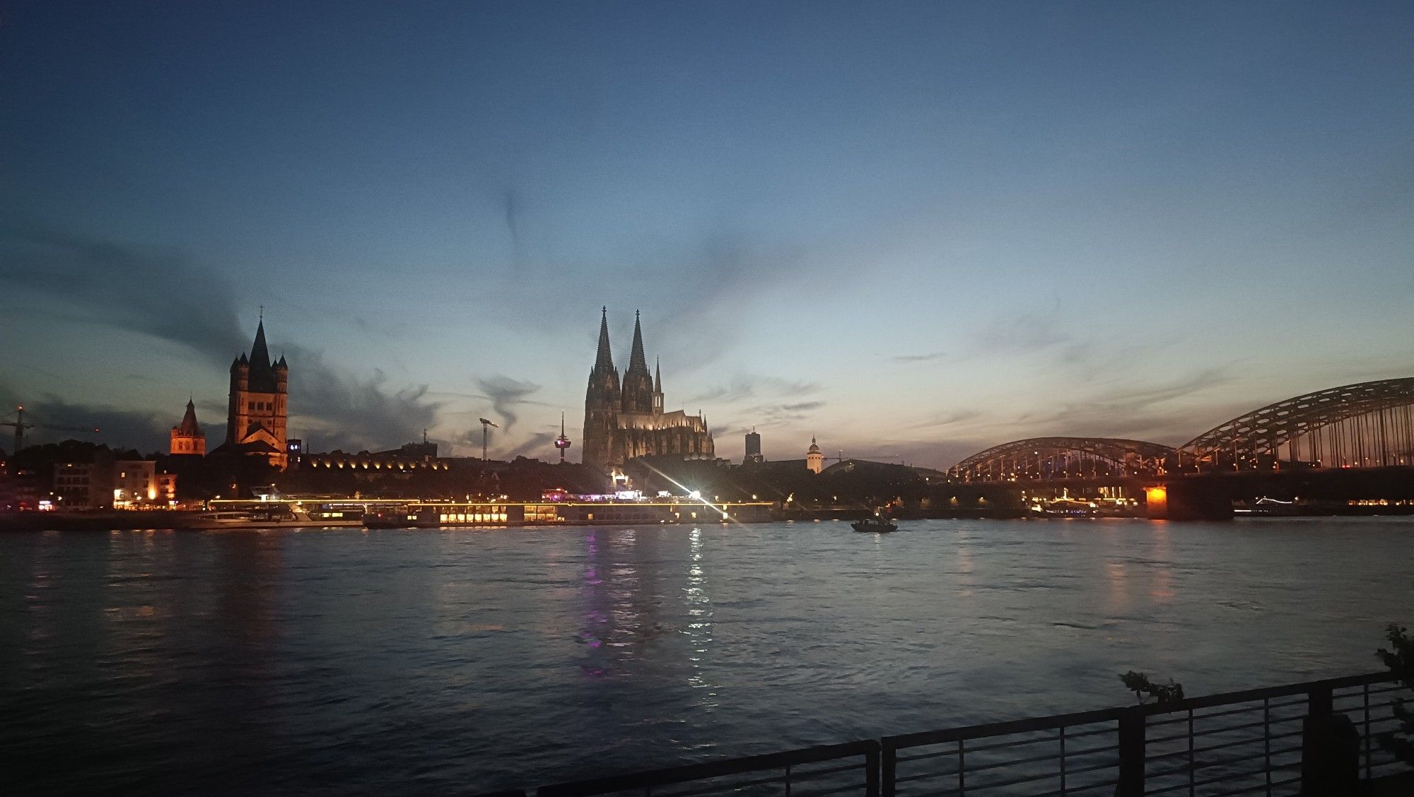 Blick vom rechten Rheinufer über den Rhein auf die Kölner Stadtsilhouette mit Groß St. Martin, Dom, Hohenzollernbrücke. Die Sonne ist untergegangen, ein paar letzte Strahlen erleuchten den Horizont.