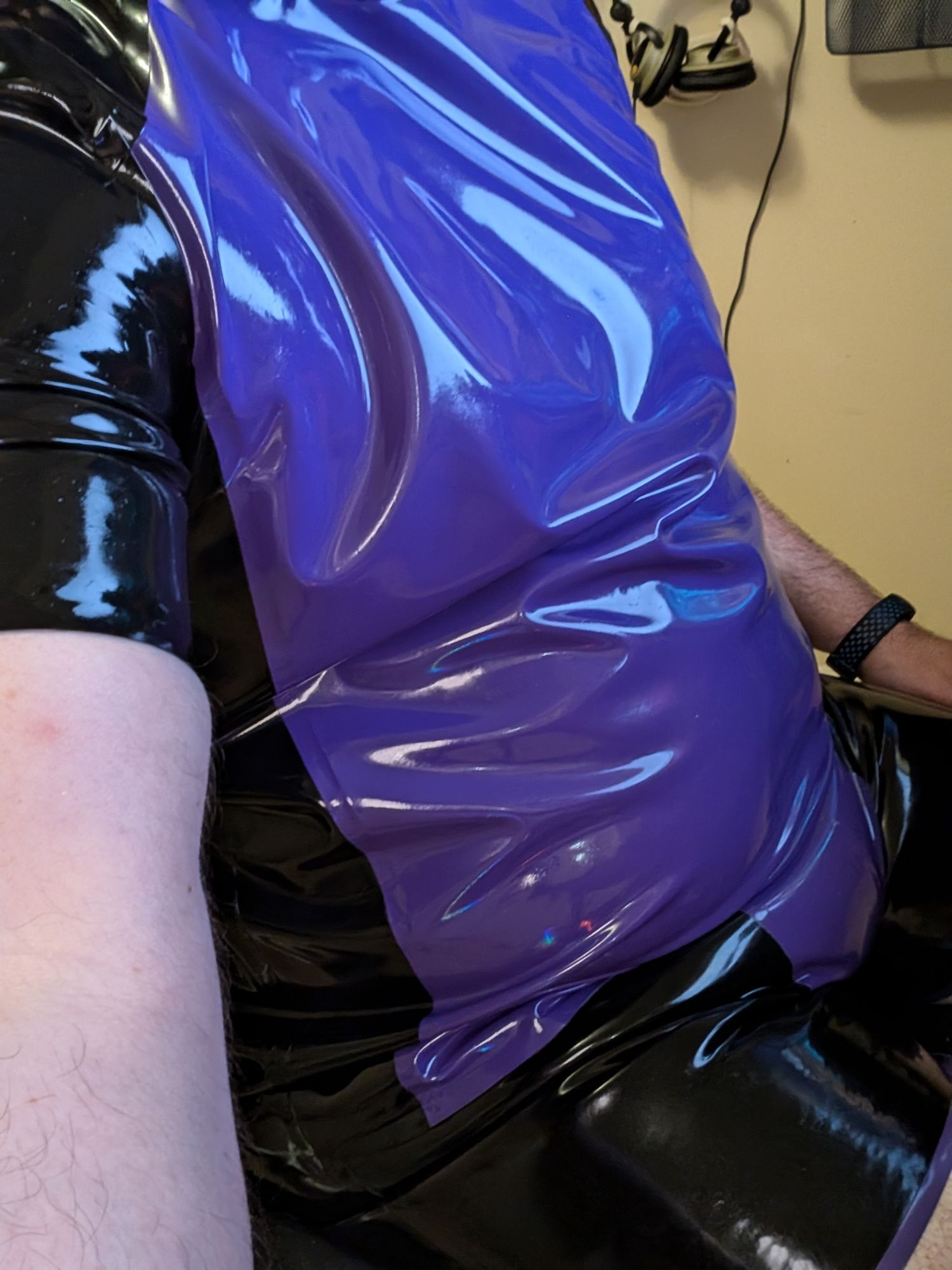 A photo of the torso and hips of a guy wearing a black latex shirt with a purple front panel, and black latex shorts with a purple crotch. He's very shiny wearing it.