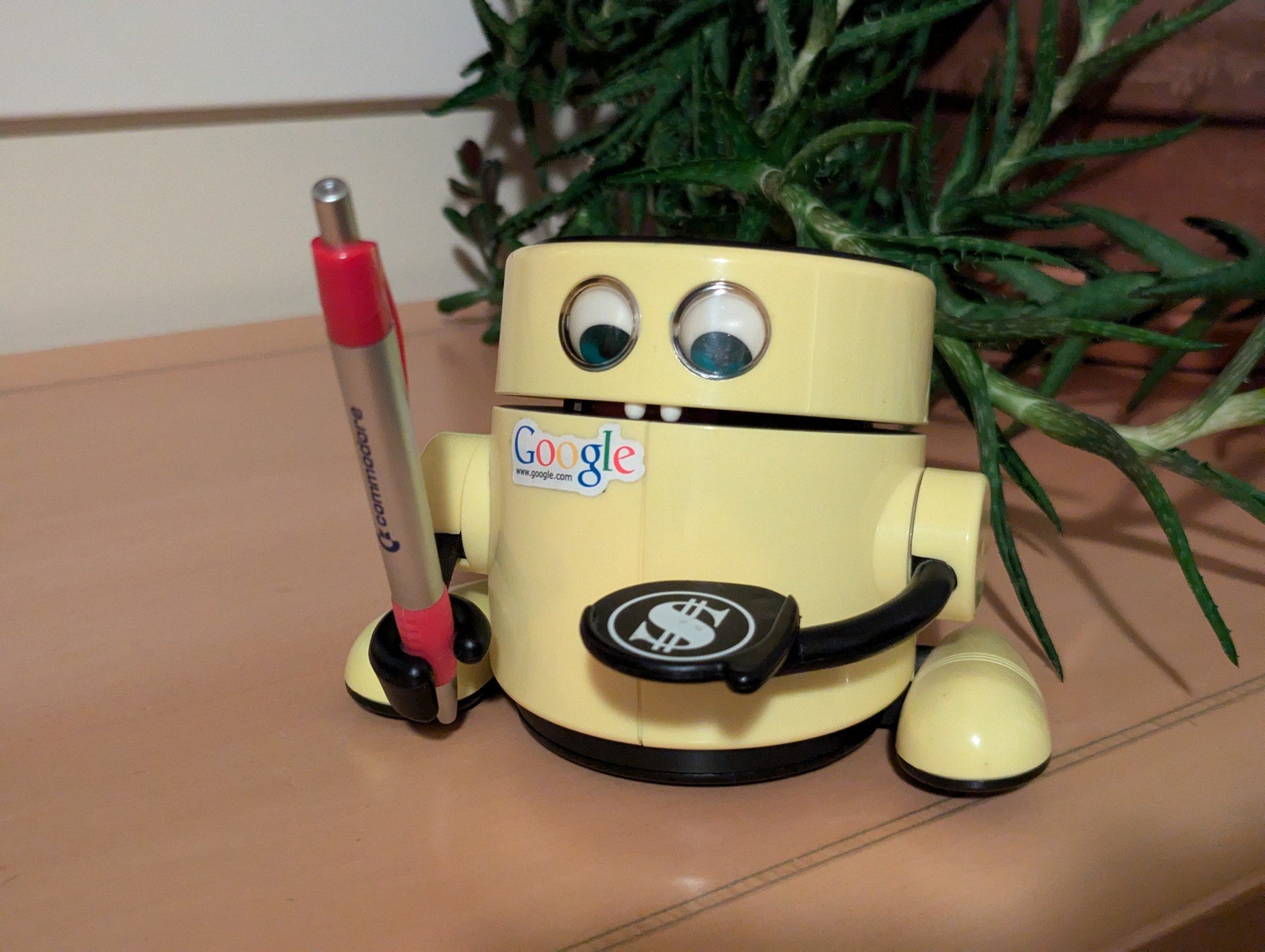 Cylindrical toy yellow robot holding a pen. It has human-like eyes and is holding a pen in its right hand. Its left "hand" is more of a tray, with a dollar sign printed on it.