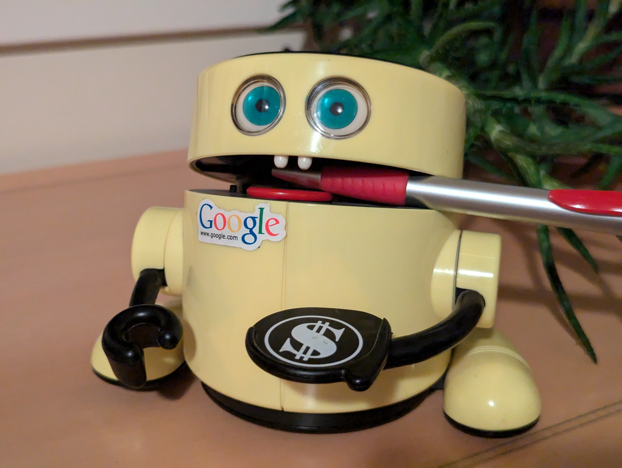 The same cylindrical toy yellow robot. The pen is now in its mouth, and which is now slightly open. Two hemispherical white "teeth" are visible above the pen, and a red plastic "tongue" is visible below the pen. The green eyes are now looking forwards, instead of slightly down.