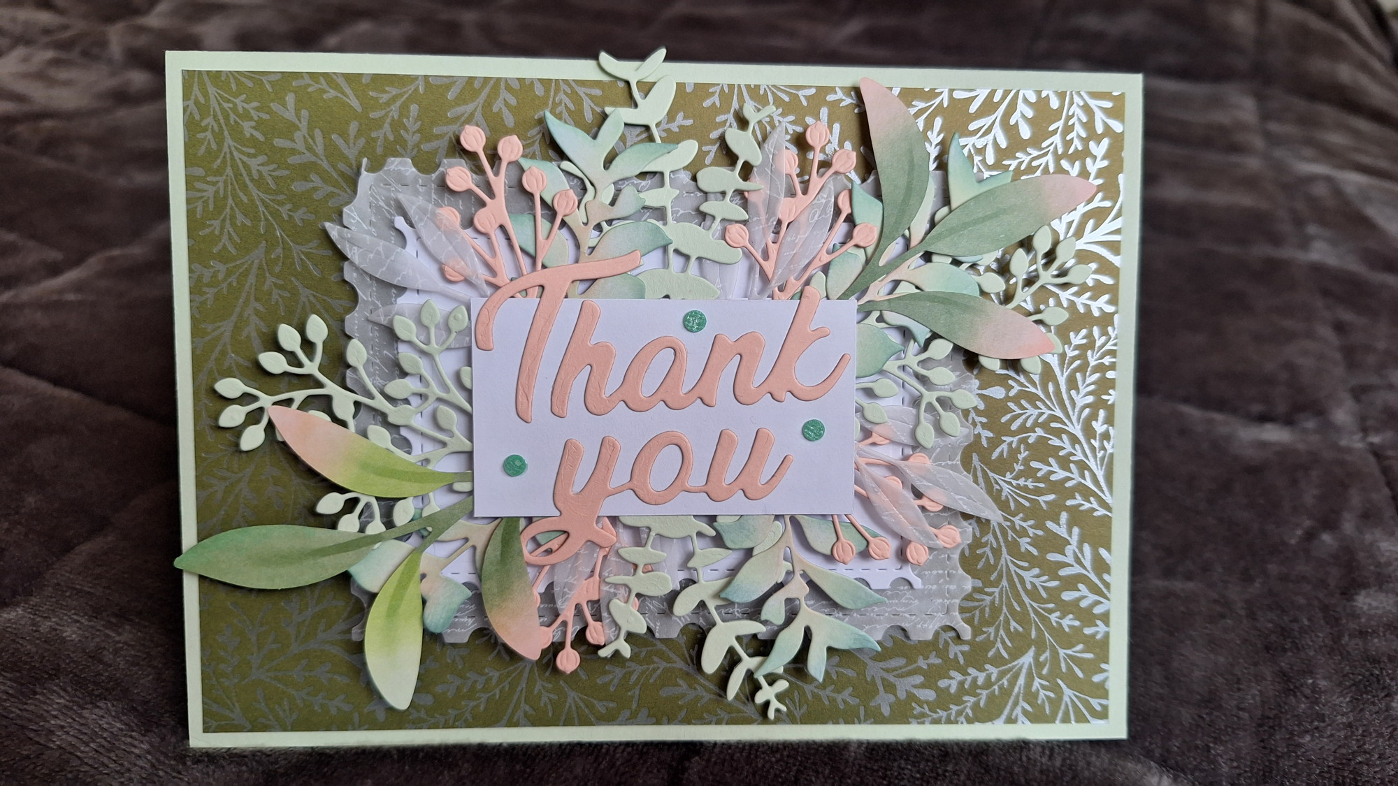 A landscape, top fold card. The base card is pastel green, layered with a metallic patterned paper that's moss green with silver foliage. A pastel pink "Thank You" sits on top of a white rectangle card, dotted with 3 green gems. The white rectangle card is surrounded with various pink, green, and white foliage die cuts.