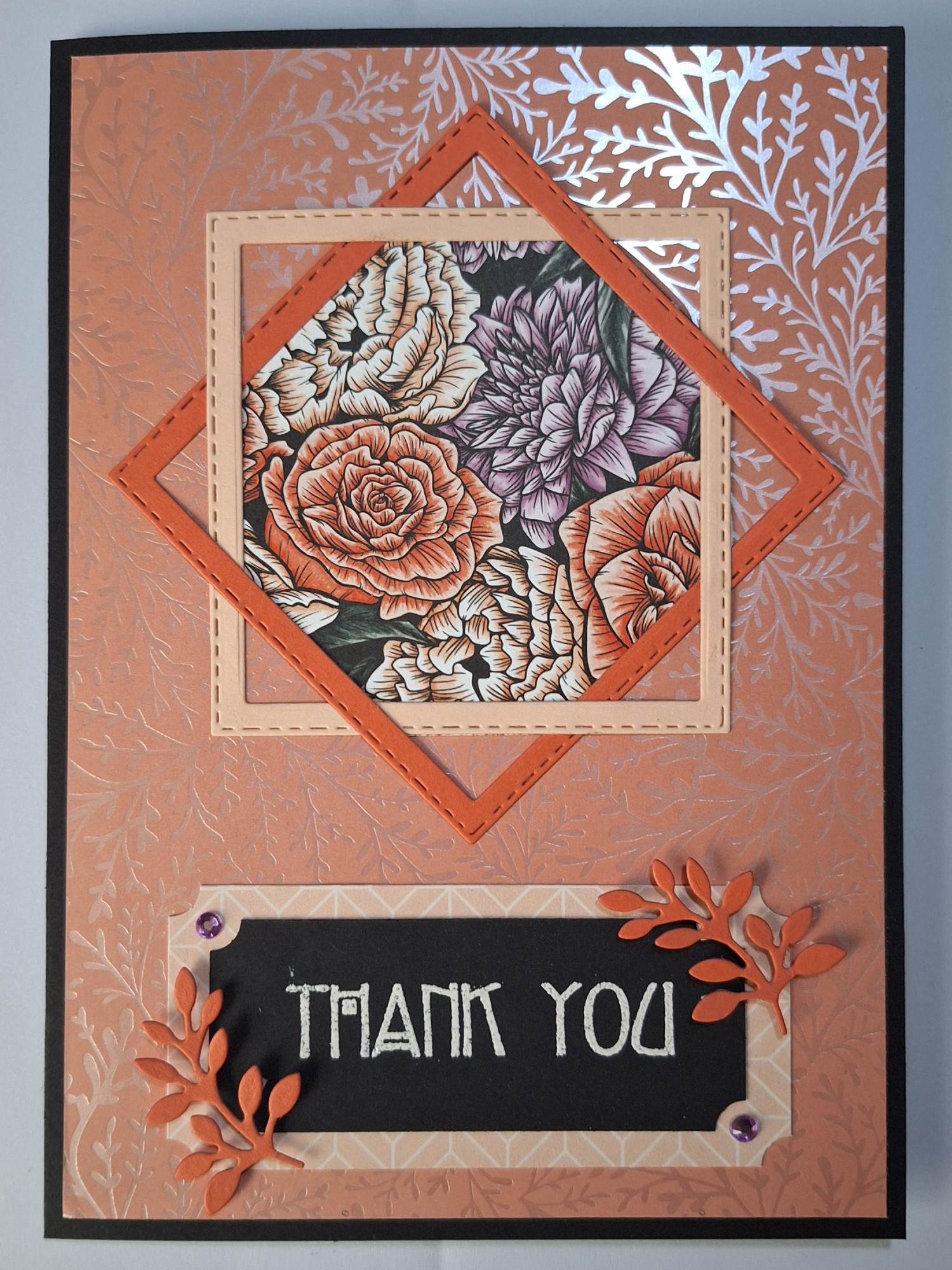 A portrait card with a black base.
Layered with a shiny peach, and silver foliage patterned card. 
2 square frames, one peach, one orange, layered on top of each other at an angle, with floral patterned paper inside them - the colours are peach, orange, purple, green, and black.
Below the squares, is a black placard, with "Thank You" with peach embossing, and orange foliage die cuts, with purple gems, framing the opposite corners.