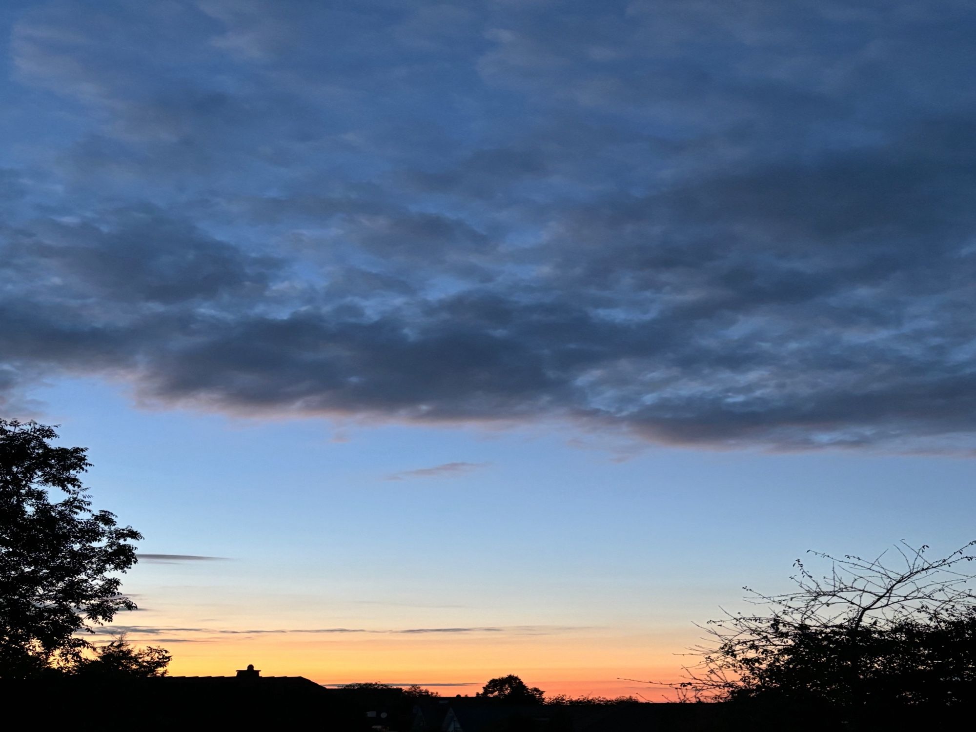 Am linken und rechten Bildrand sind die Silhouetten von Bäumen zu erahnen. Im Hintergrund leuchtet nachtblau dunkel der ab der Mitte nach oben etwas bewölkte Himmel im Hauch zwischen hell und ins dunkle Blau gehend. Die Sonne schickt Strahlen über den Himmel bis zu den Wolken und beleuchtet den unteren Himmel sanft orange, ist selbst jedoch nicht mehr zu sehen.