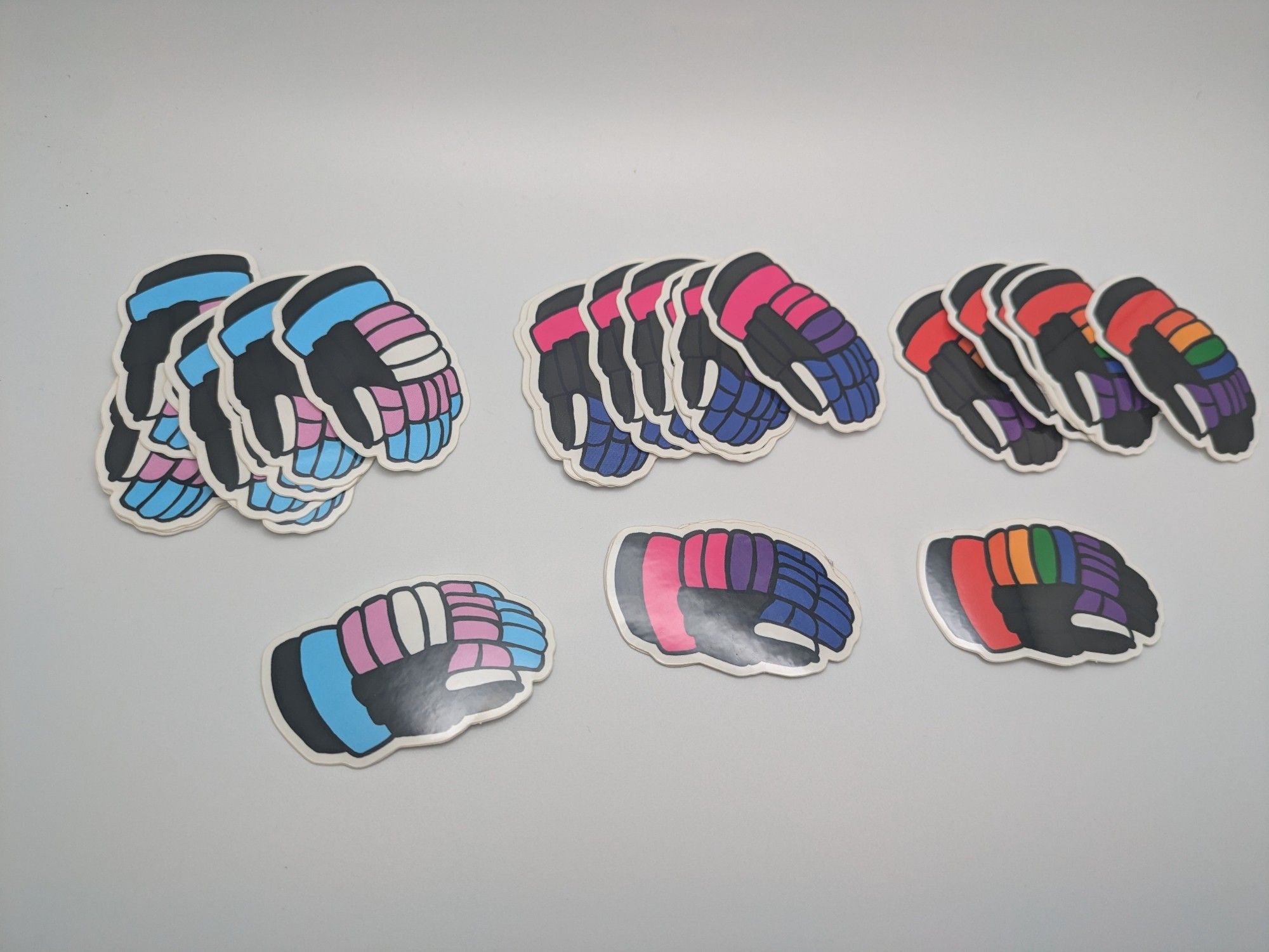 A bunch of stickers shaped like hockey gloves, some colored for trans pride, some for bi pride, and some rainbow.