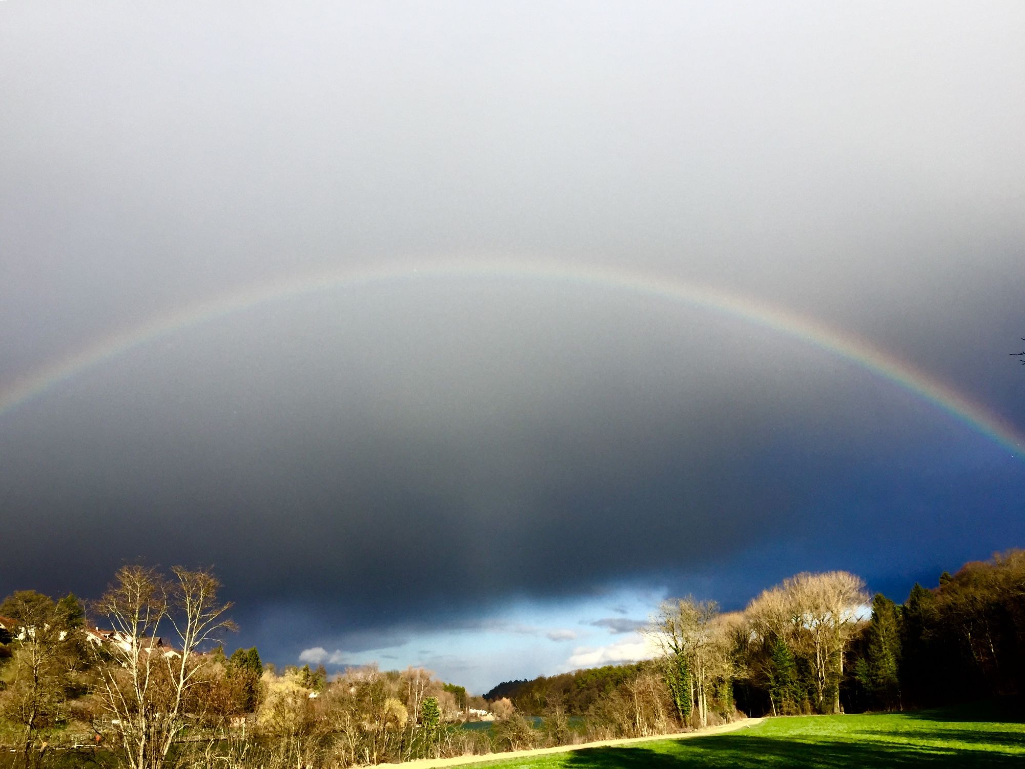 Ein Regenbogen vor dunkeln Wolken. Darunter im Sonnenlicht Bäume und ein grünes Feld.