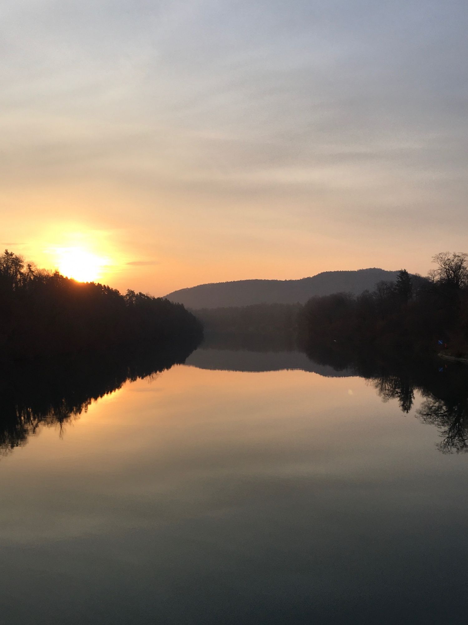 Aussicht von einer Brücke flussaufwärts. Die Wasseroberfläche ist ruhig. Beidseitig sind die Ufer von Wald gesäumt. Links geht die Sonne auf und  färbt den Himmel zart orange.