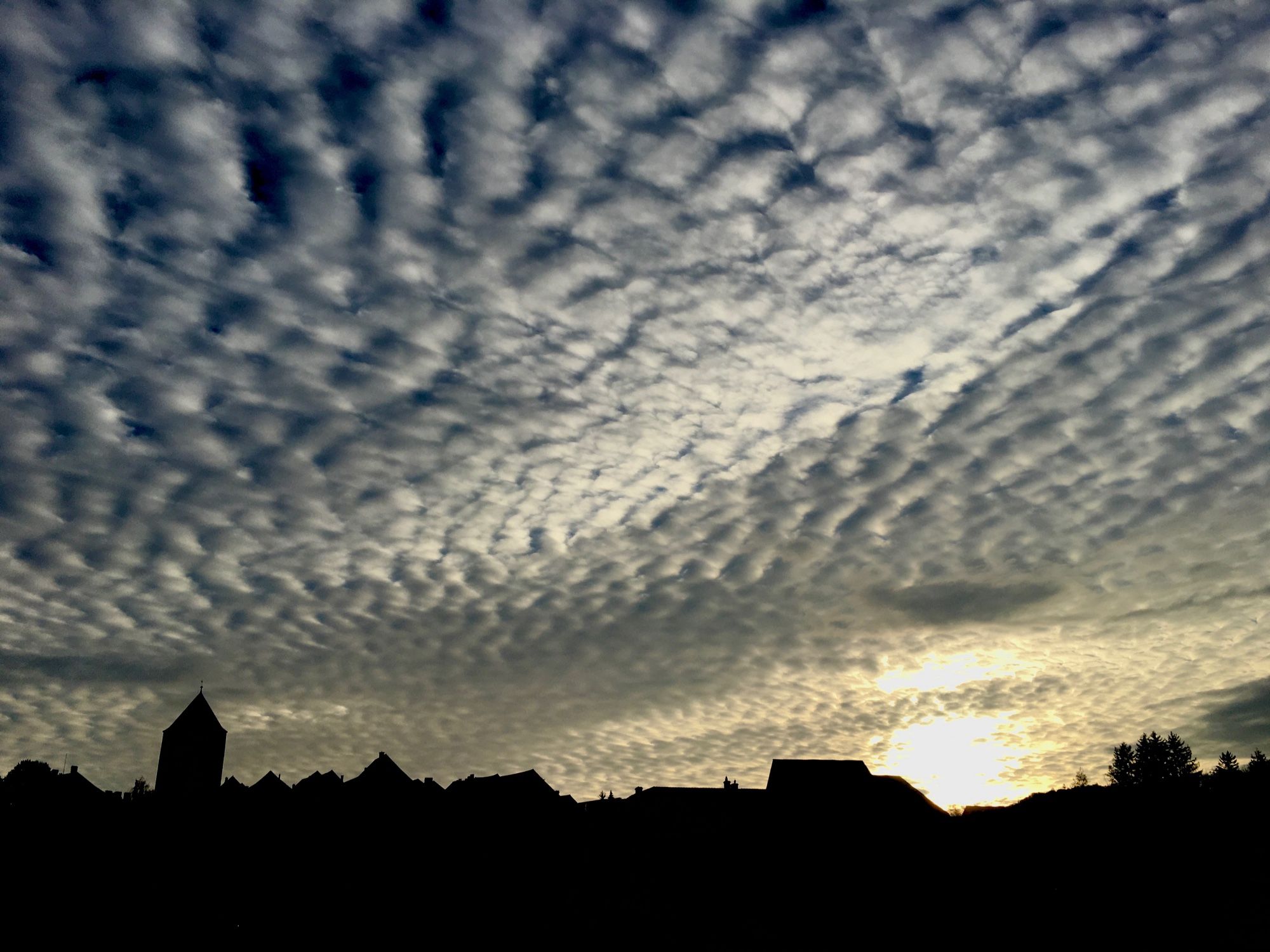 Abendhimmel, die Sonne verschwindet hinter der schwarzen Silhouette einer Gebäudereihe am Horizont. Der Himmel ist von Wolken in schmalen, parallel verlaufenden Streifen bedeckt.