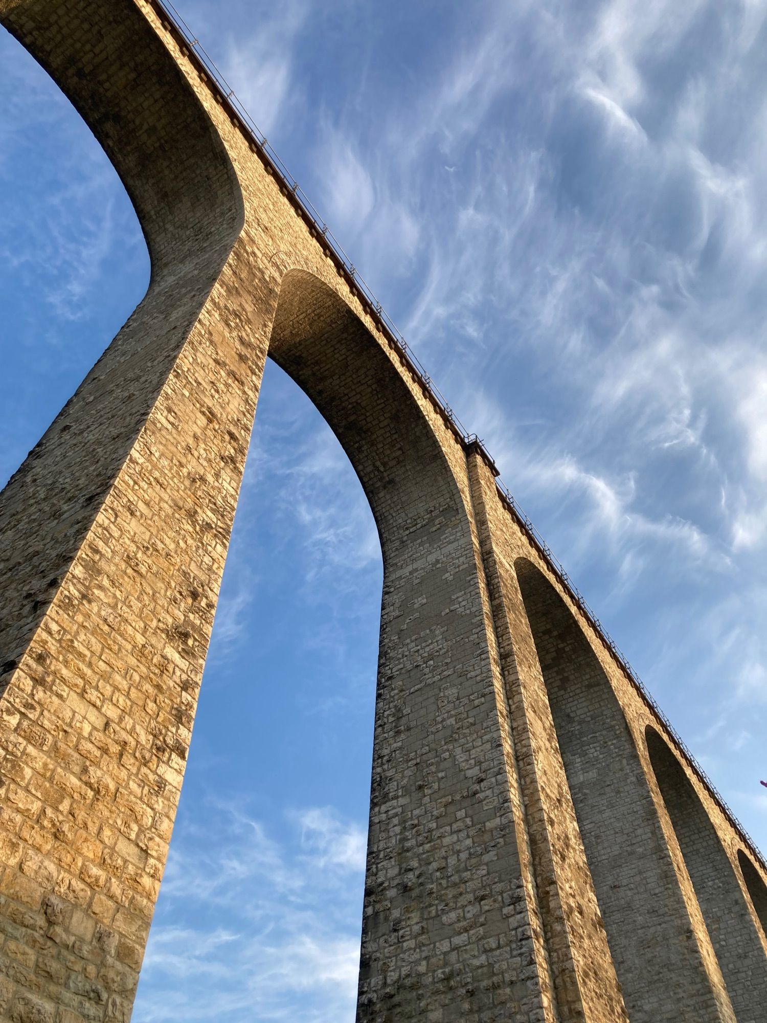 Looking up to a tall stone brick railway bridge from below the arches. It’s lit up by the light of the rising sun. Blue sky with wispy cirrus clouds.