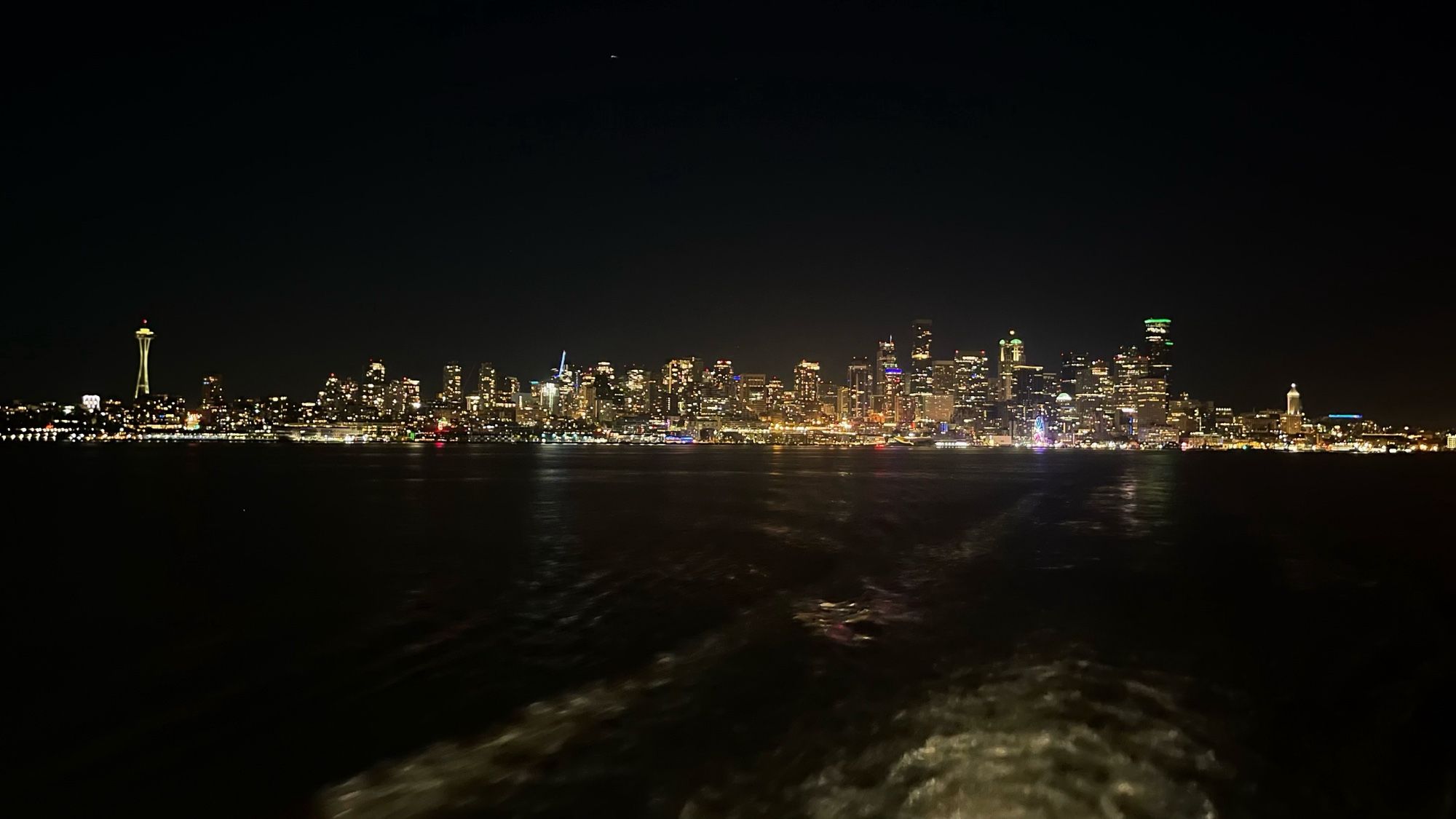 A night shot of downtown Seattle from the aft pickleforks of the MV Chimacum. The water is almost black, except for the white turbulence turned up by the propeller. The Space Needle is at far left, and the waterfront is well lit all the way south past Colman Dock.