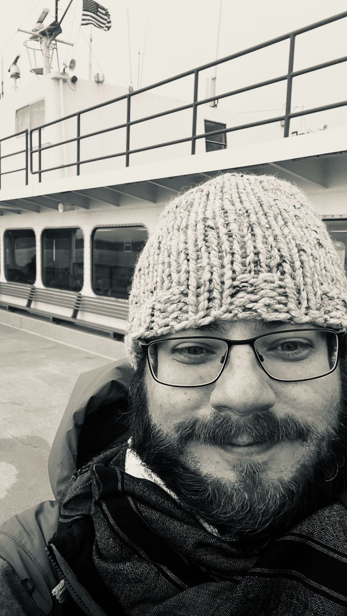 A black and white photo of a bearded gentleman standing on the weather deck of the MV Kaleetan. He is wearing a crocheted beanie, a scarf, and a heavy puffer jacket. A row of benches and windows are in the background with an American flag waving from the mast in the upper lefthand corner.