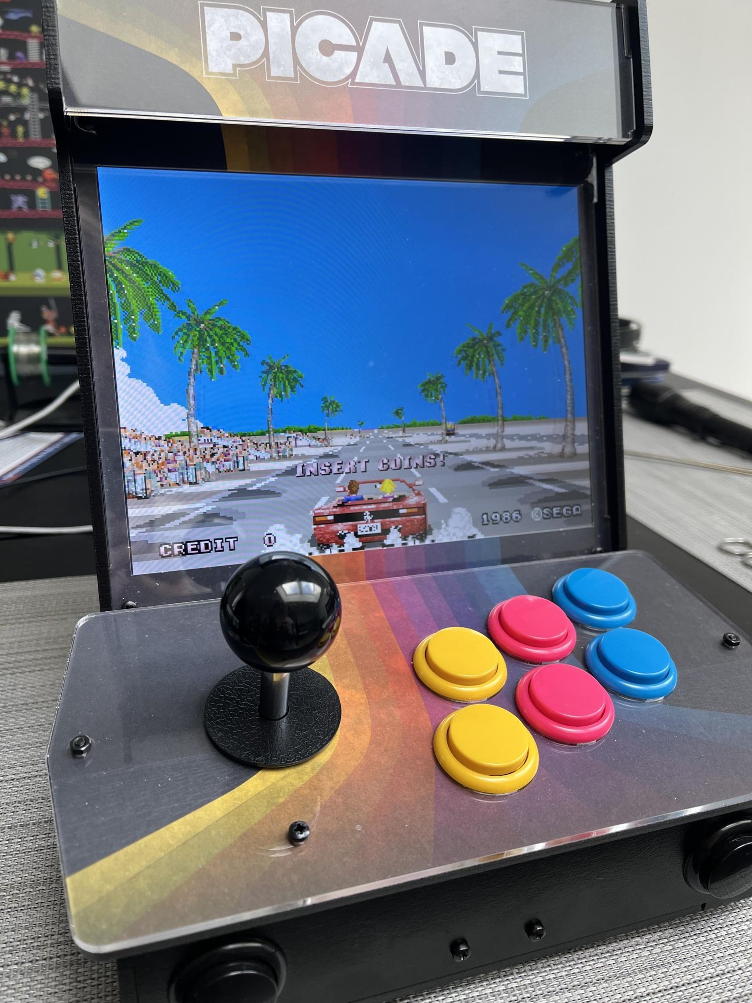 A small “picade” arcade game console with a joystick, 6 coloured buttons and a screen. It is playing Outrun