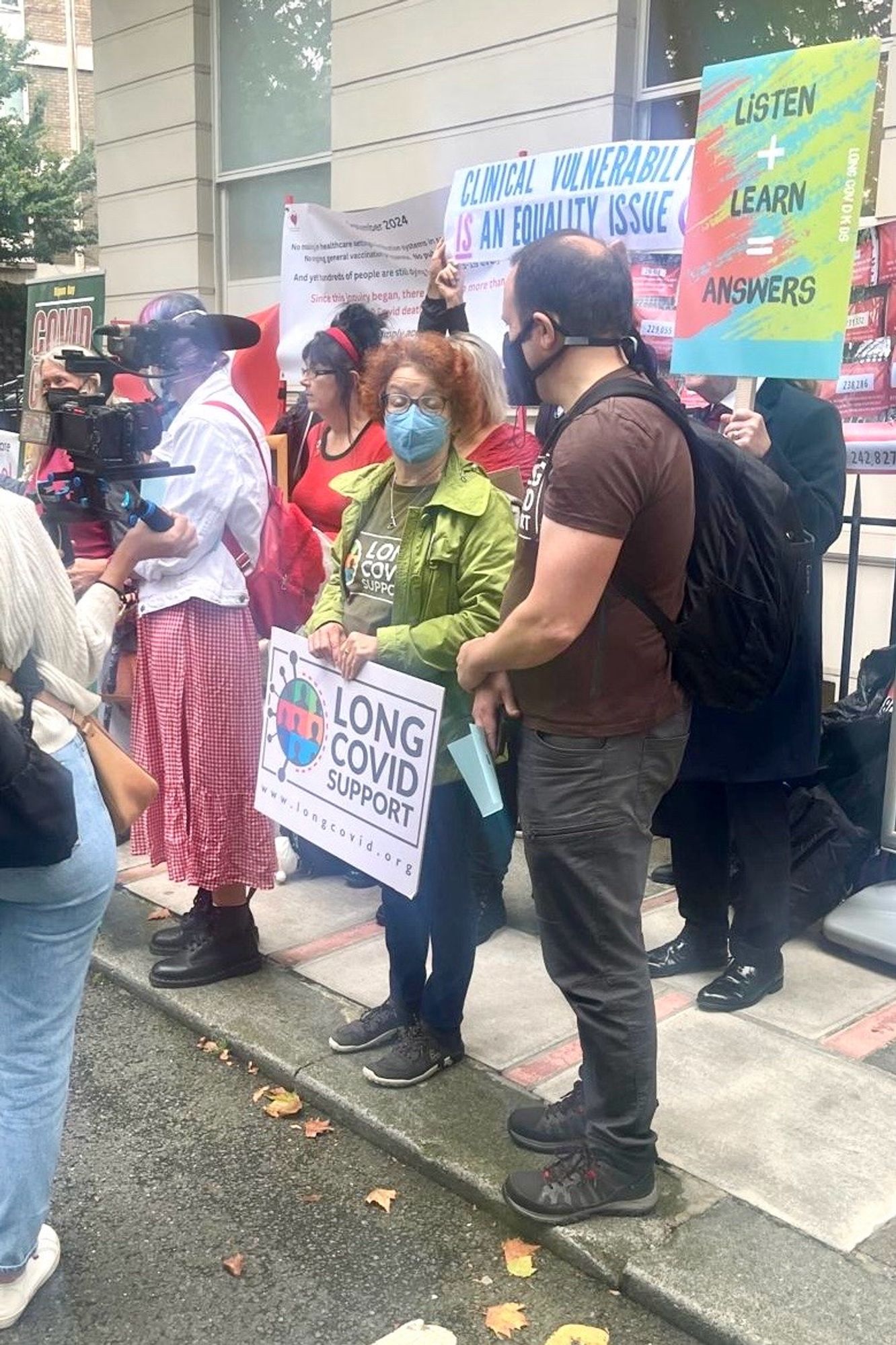 Photo of a group of people stood outside the Covid Inquiry including a man and woman both wearing Long Covid Support tops. The woman is holding a placard bearing the Long Covid Support logo and website address www.longcovid.org. There is a woman facing them holding a camera with microphone.