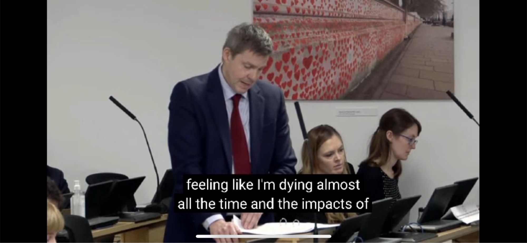 Man wearing jacket, shirt and tie reading from documents on a desk. There is a microphone in front of him. Subtitles read “feeling like I’m dying almost all of the time and the impacts of”.