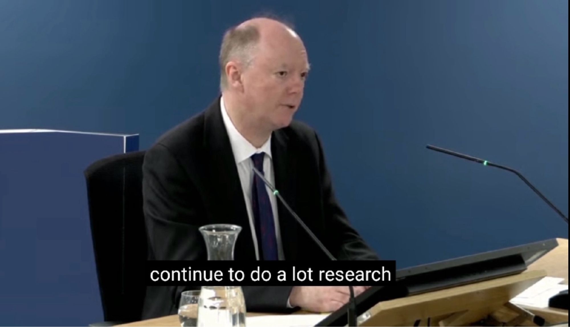 Man wearing shirt, jacket and tie sat behind a desk with a microphone in front of him and a jug of water to his right. Subtitles read “continue to do a lot research”.