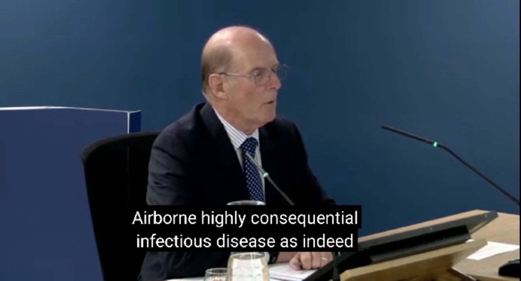 Dr. Barry Jones wearing a shirt, dark jacket and tie, sat behind a desk with a microphone in front of him. Subtitles read “Airborne highly consequential infectious disease as indeed”.