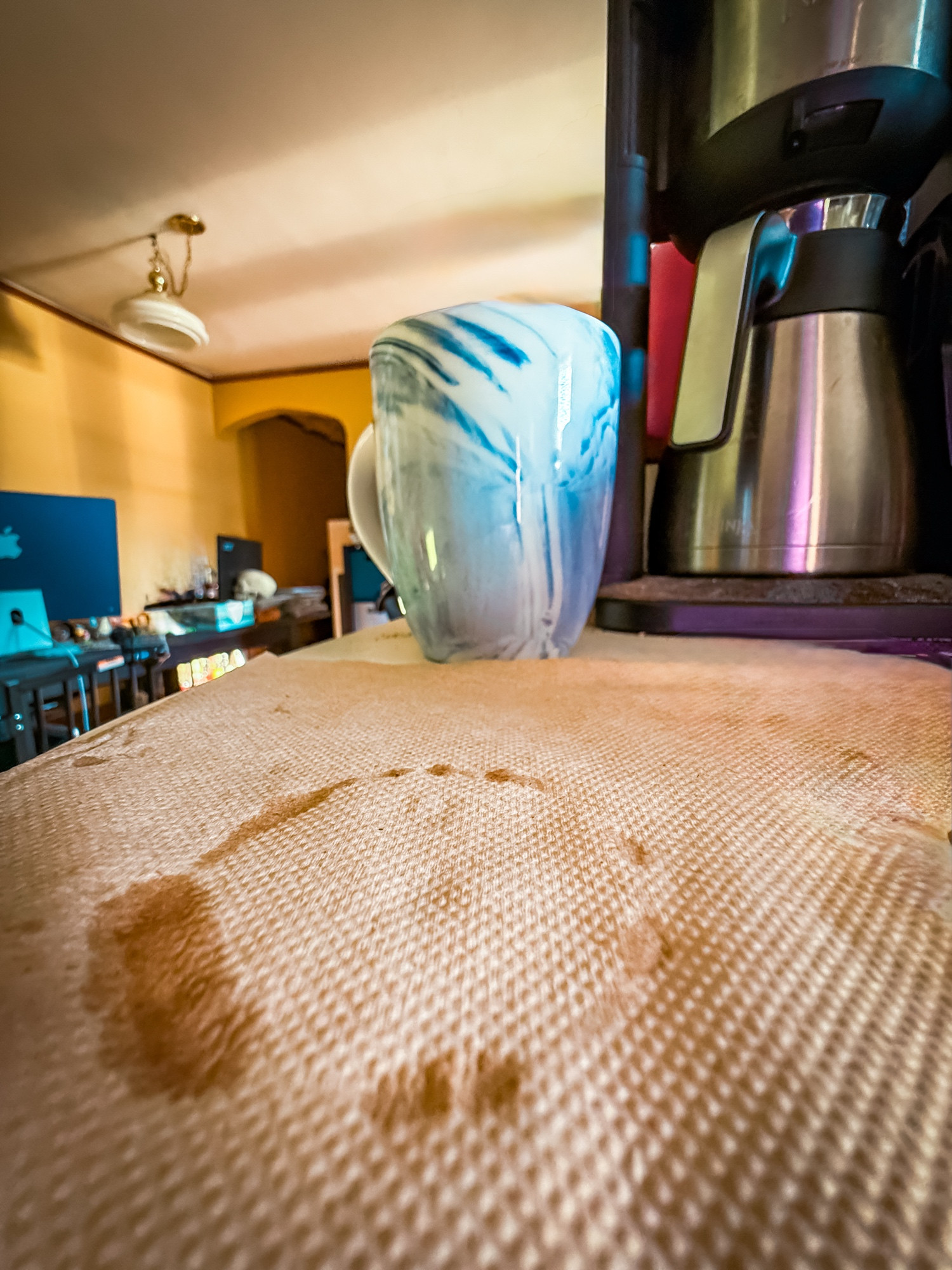 Image of a coffee mug and coffee maker.  The foreground is a brown napkin with a wet circular print from the bottom of a mug.