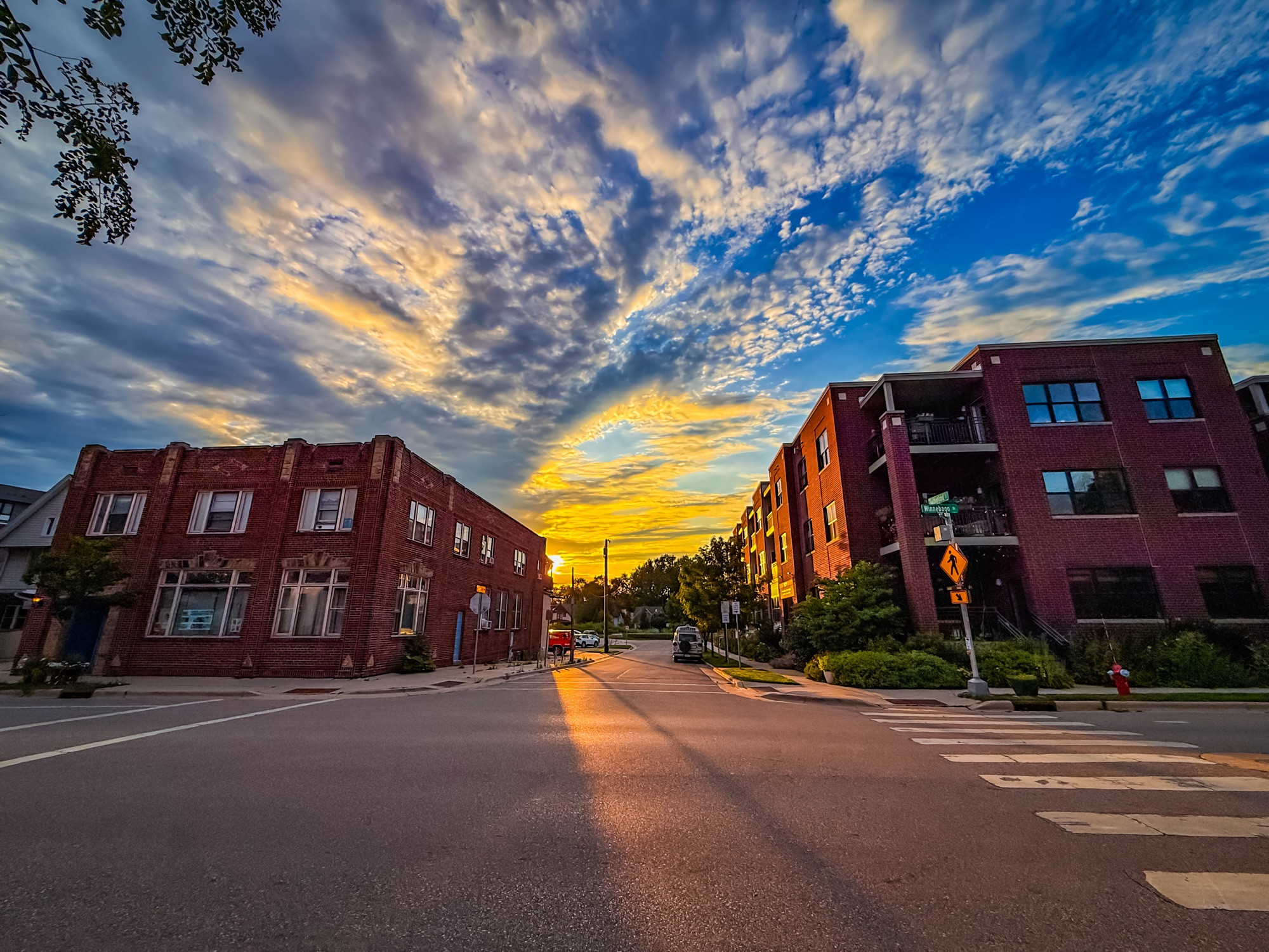 Image of a sunset between two red brick buildings.  The sky above is full of dynamic cloud formations and a shadow line cuts down the street in the foreground.