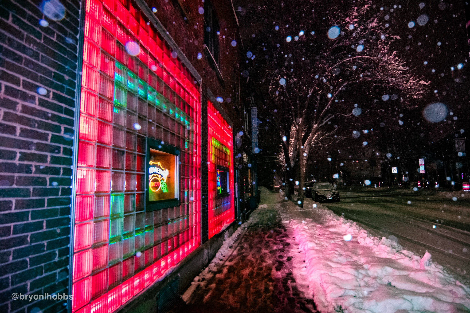Image of a brick wall with a “window” of glass blocks lit up by neon lights at a side angle during snowfall