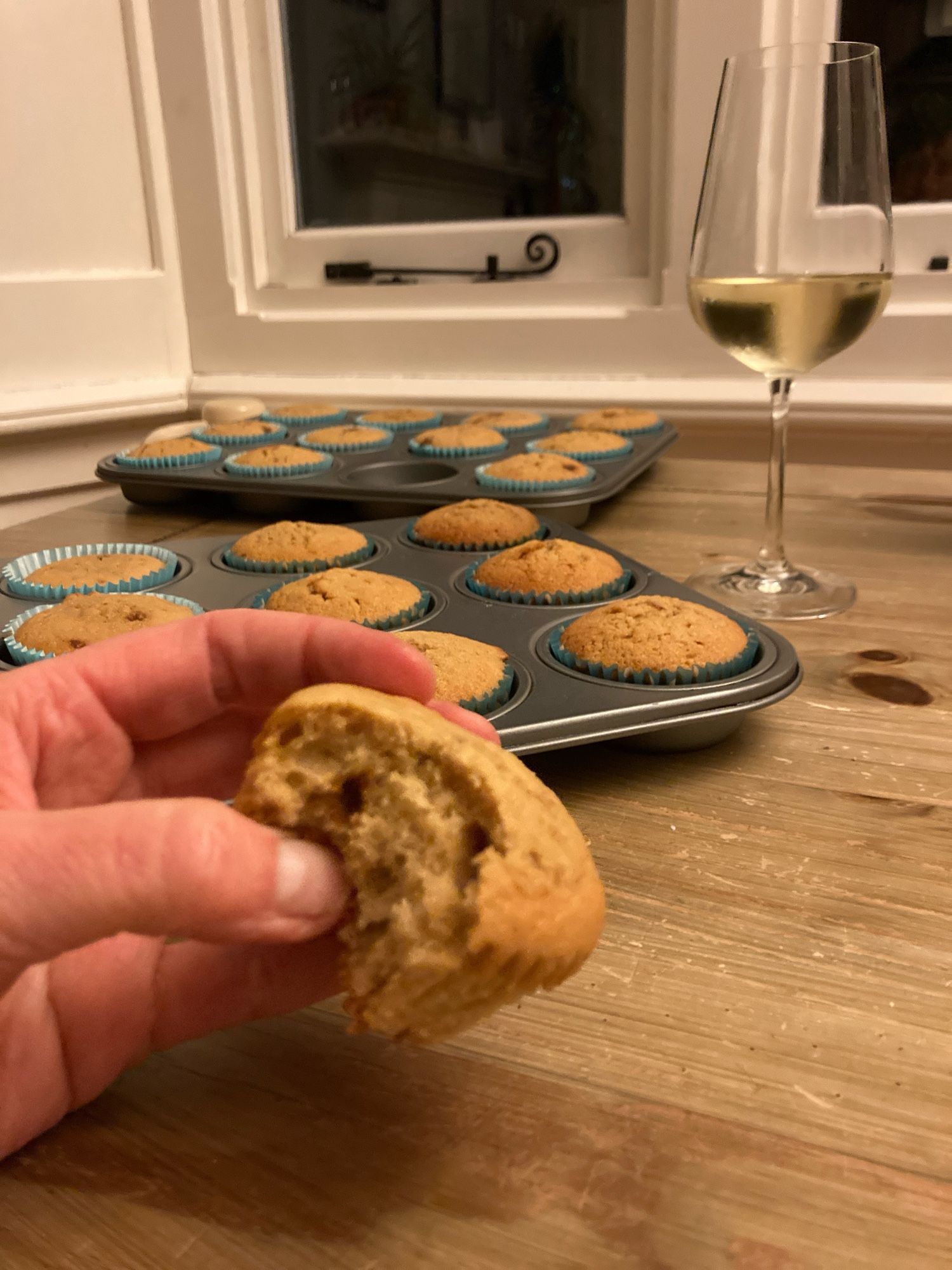 Half eaten cup cake held in front of tray with more similar cakes. Also a glass of white wine