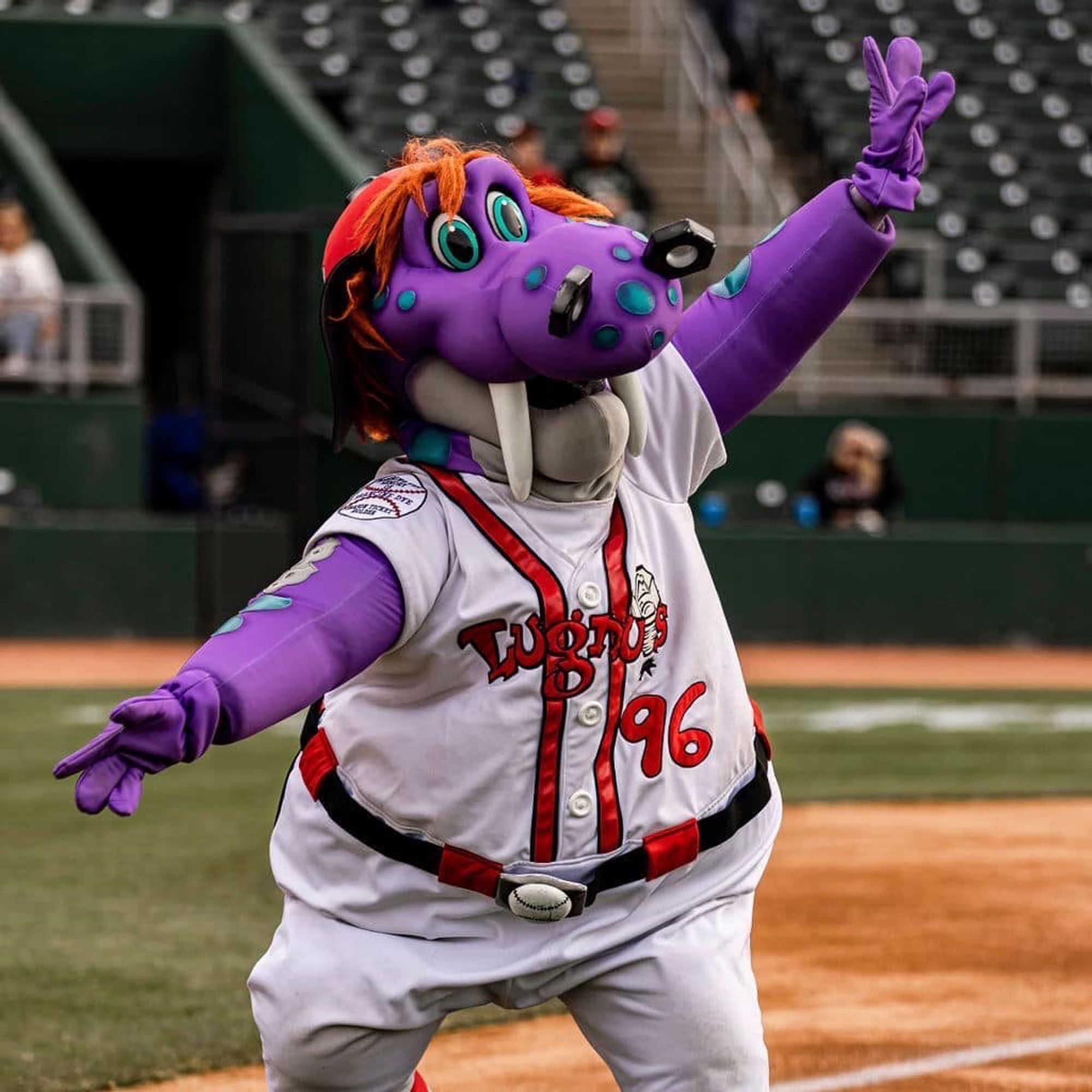 Big purple dragon(?) with blue spots and lugnuts for nostrils, wearing a baseball uniform and posing on a baseball field.