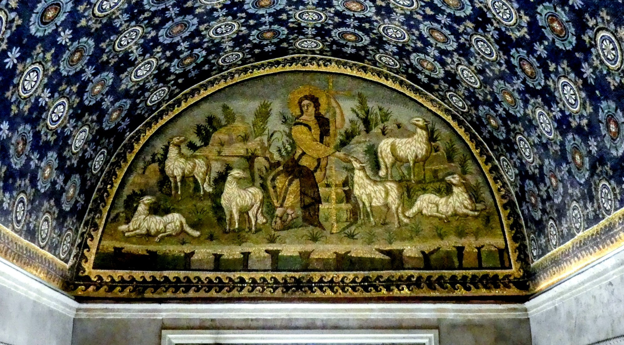 Another interior mosaic from the Mausoleum of Galla Placidia. The arched ceiling is adorned with a deep blue mosaic studded with golden stars and white floral patterns. Below, a lunette mosaic shows Christ as the Good Shepherd, surrounded by sheep in a pastoral setting. The figure of Christ has a golden halo and is dressed in golden robes, standing among trees and rocks.