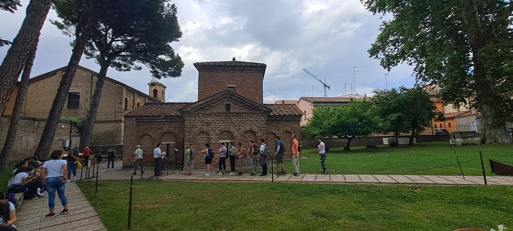 Exterior view of the Mausoleum of Galla Placidia in Ravenna, Italy. A small brick building with arched windows and a central tower, surrounded by trees. A group of tourists is lined up outside the entrance, waiting to enter. The sky is overcast, and modern buildings are visible in the background.