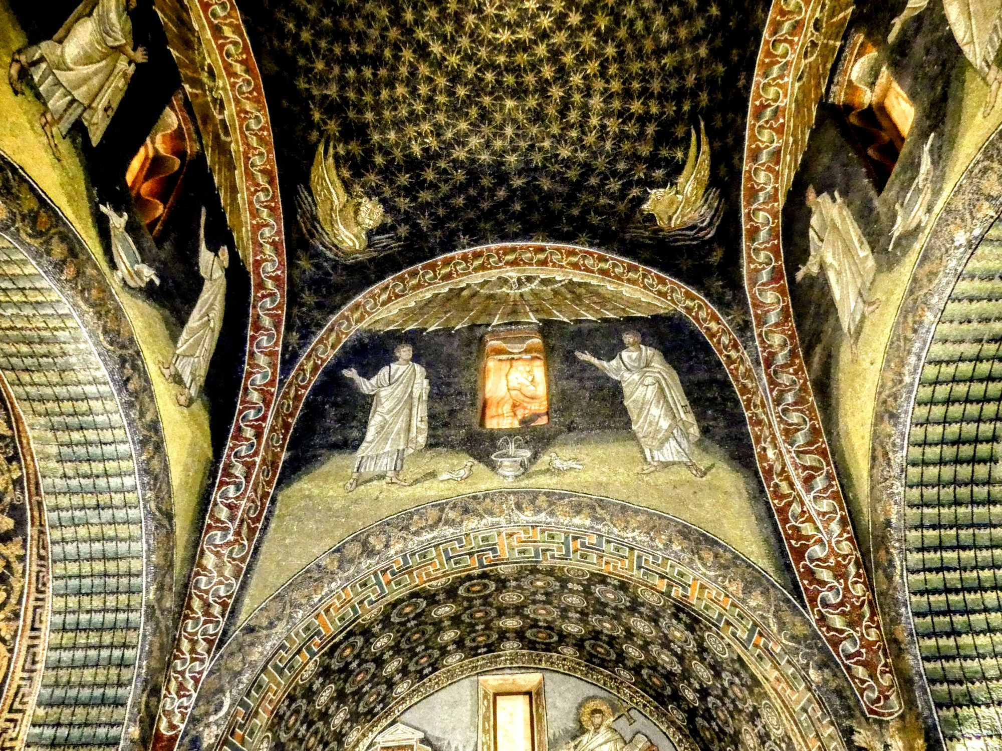 Interior of the Mausoleum of Galla Placidia in Ravenna, showing intricate mosaic work. The vaulted ceiling is adorned with a dark blue mosaic studded with golden stars. A central lunette depicts two figures in white robes gesturing towards a glowing window. The arches are decorated with ornate patterns and figures. Below, another arch partially visible shows a figure holding a cross. The entire scene is richly decorated with geometric and floral motifs in gold, blue, and earthy tones.