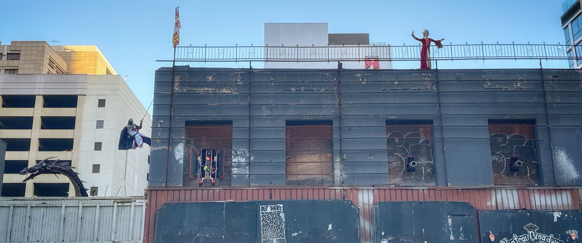 DTLA display of life-size knight climbing a building as if it were a castle with a dragon behind him, a damsel in distress on the roof and a jester sitting in a window.