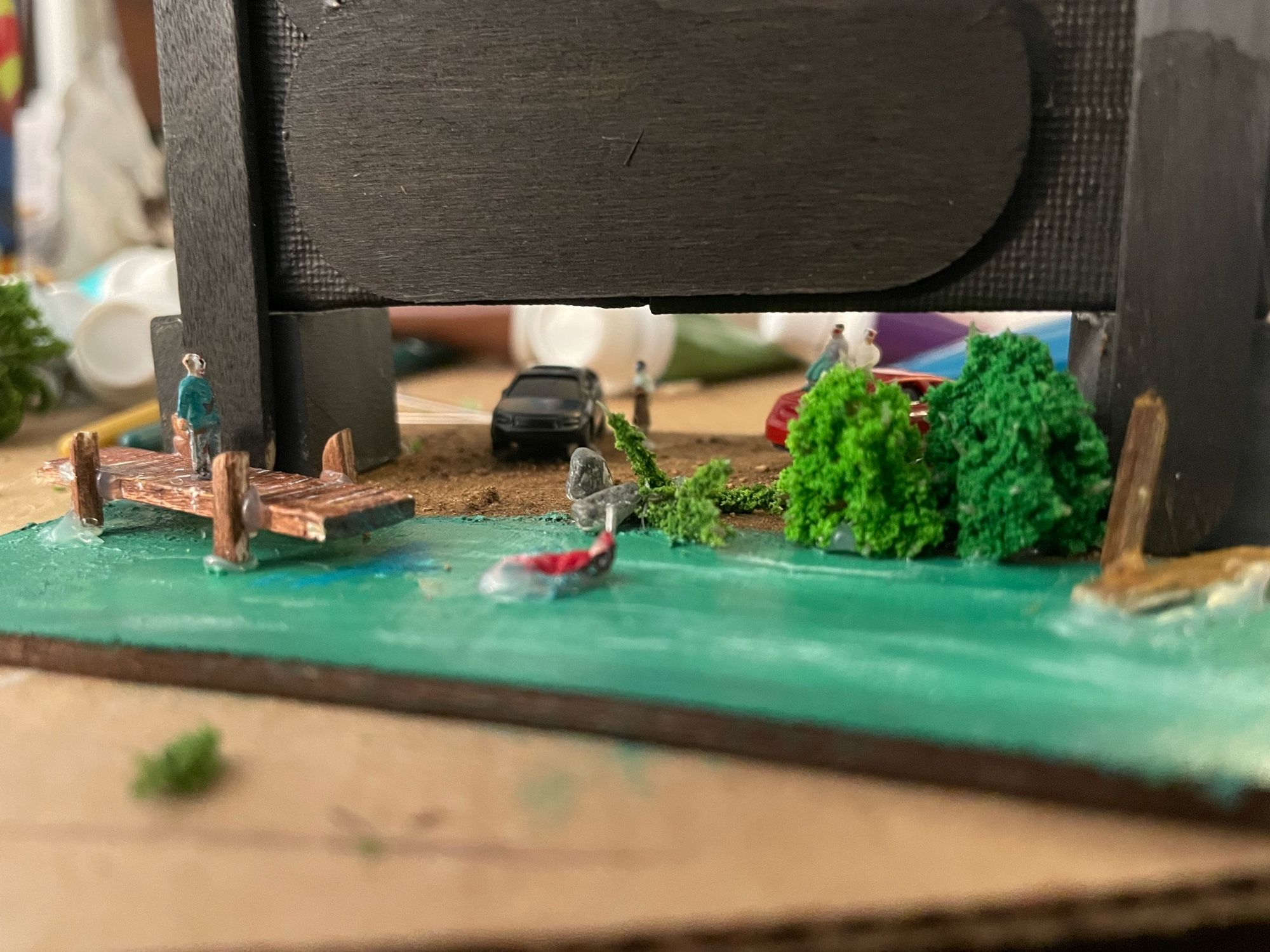 View from the back side of the tiny drive in model. A small lake scene is seen with a dock coming out into the water. A tiny masked figure is standing on the dock. In the water is a person floating with a small metal object protruding from the head area. Meant to depict a scene from Friday the 13th part 3.