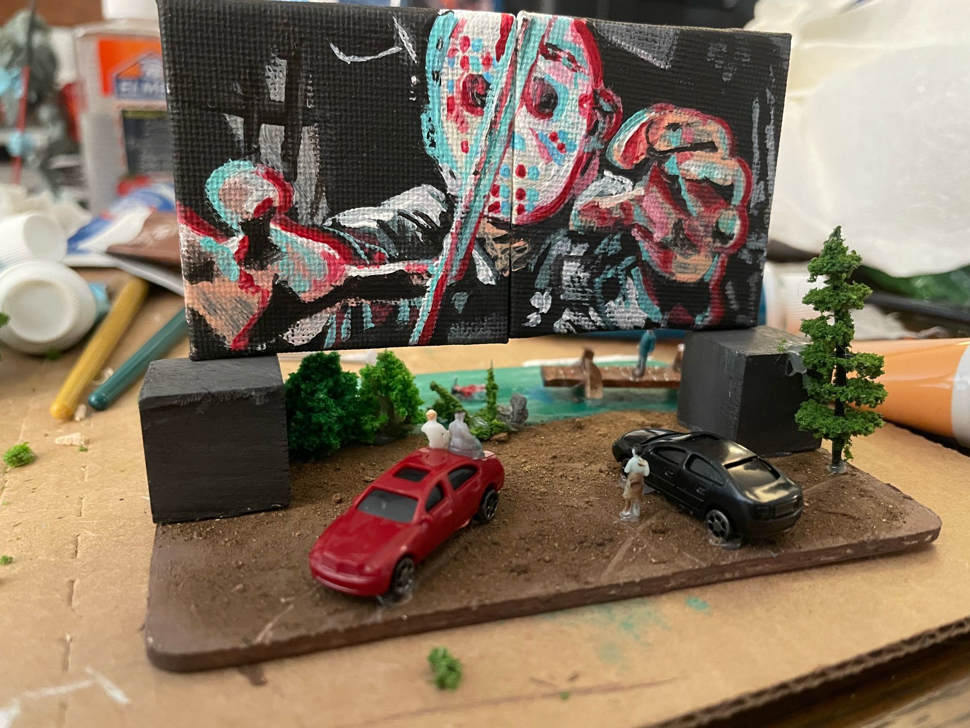 Tiny Drive-In model. Screen is hand painted mini canvas depicting Jason from Friday the 13th part 3. Small cars are parked in front of the screen in the dirt. A couple tiny people are sitting on the car. A lake scene is seen in the background behind the screen.