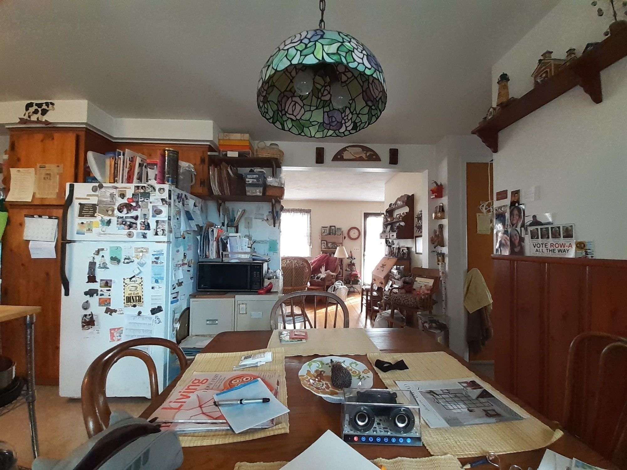 A photograph of a bright, and very lived in kitchen nook. Fridge covered in notes and magnets, a stained glass lamp hangs over the table. It was one of my favorite spaces in her home.